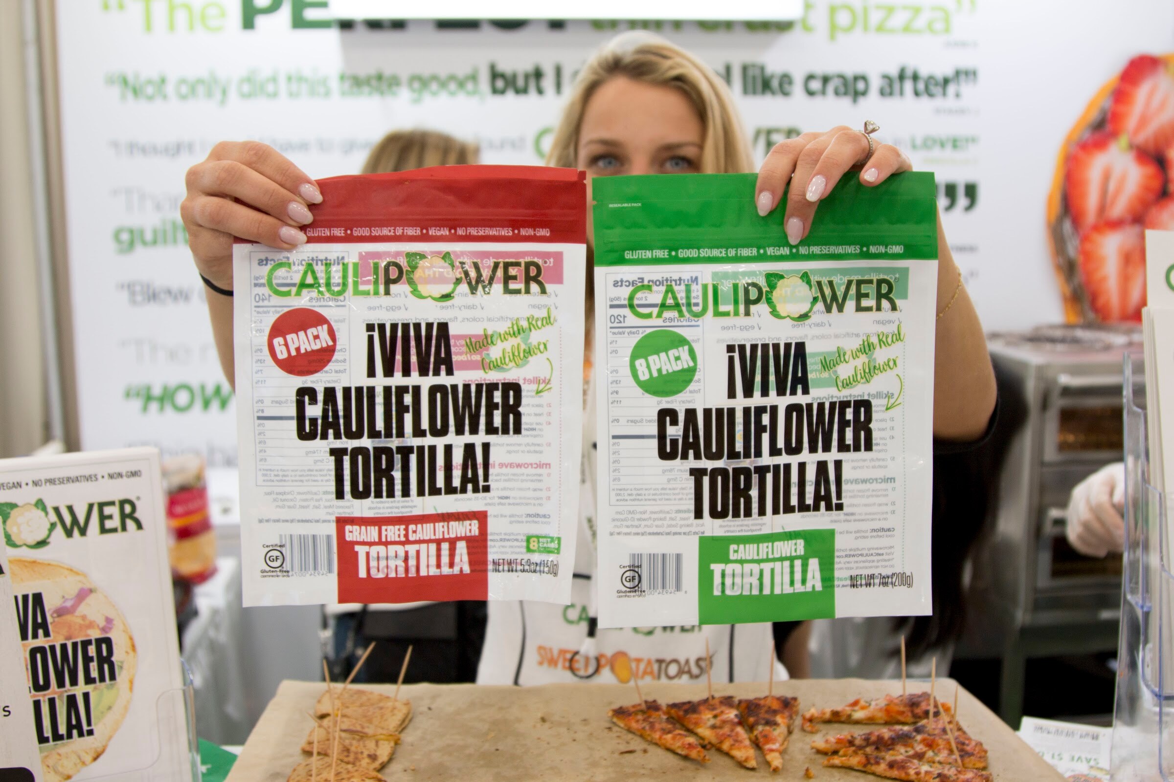 A smiling woman peeking over two Caulipower tortilla bags, one red and one green, with sample slices on a tray below.