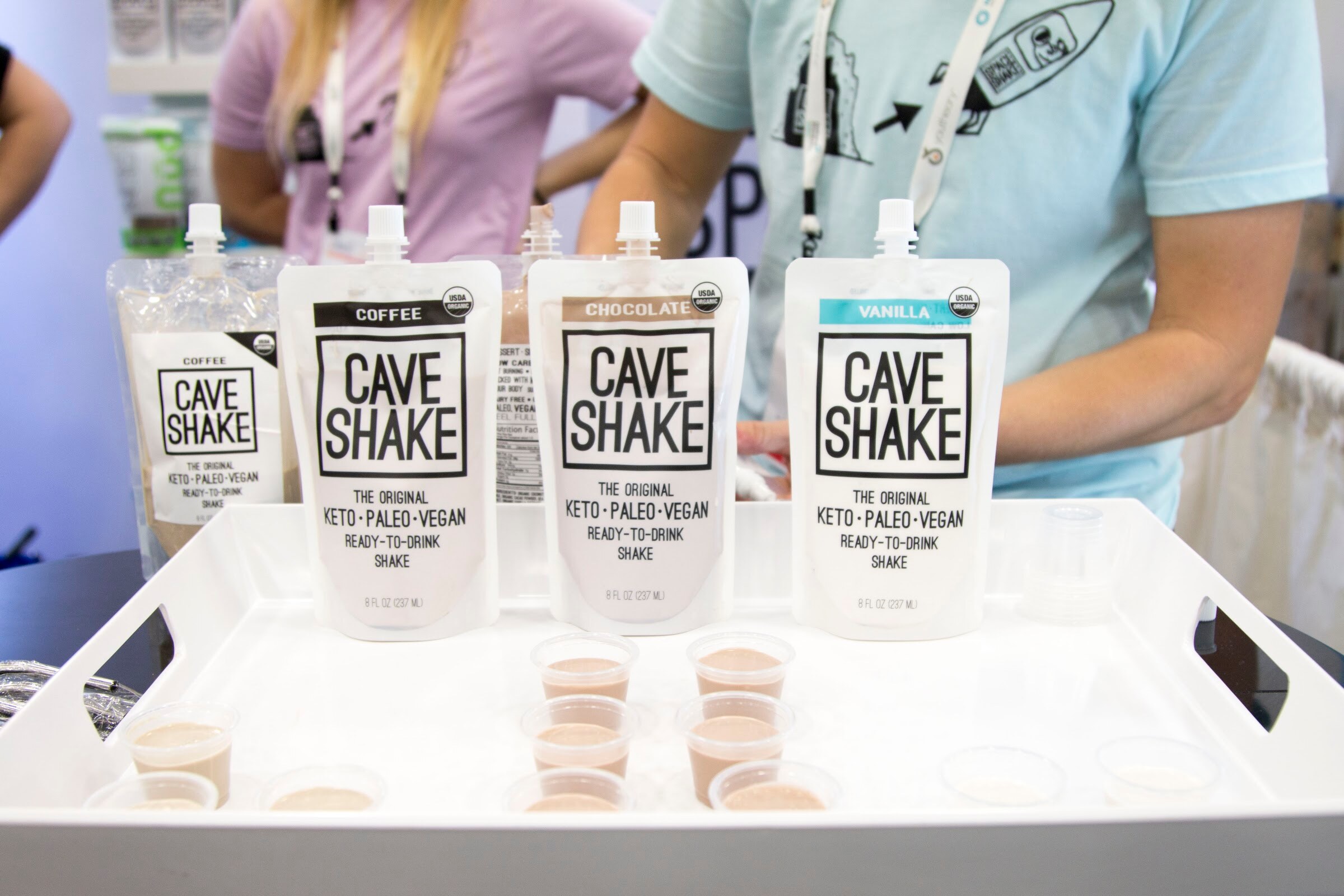 Three Cave Shake pouches on a white tray with small sample cups and two staffers standing behind.
