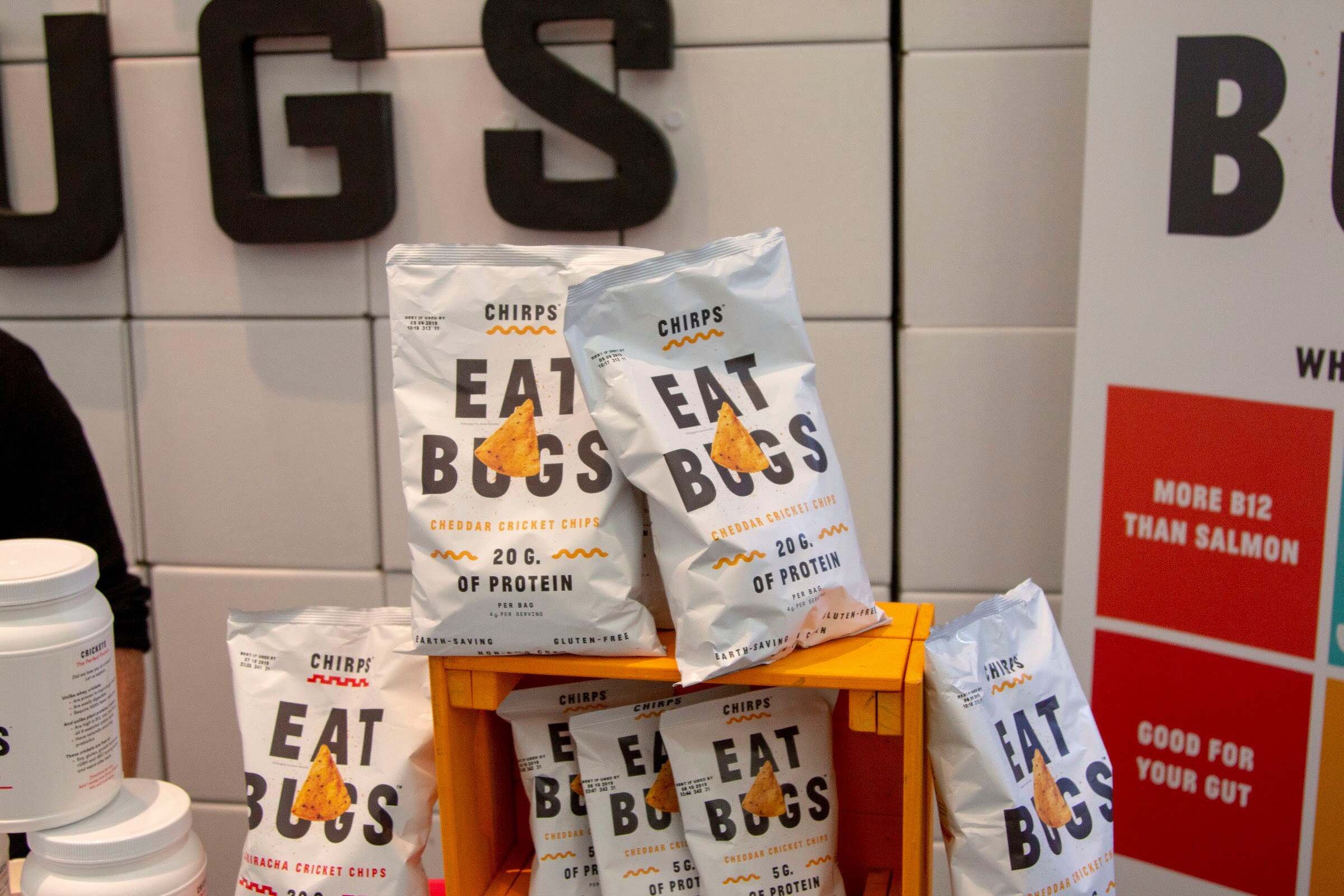 White Chirps bags printed with Eat Bugs on an orange crate against a wall of white tiles with large black letters.