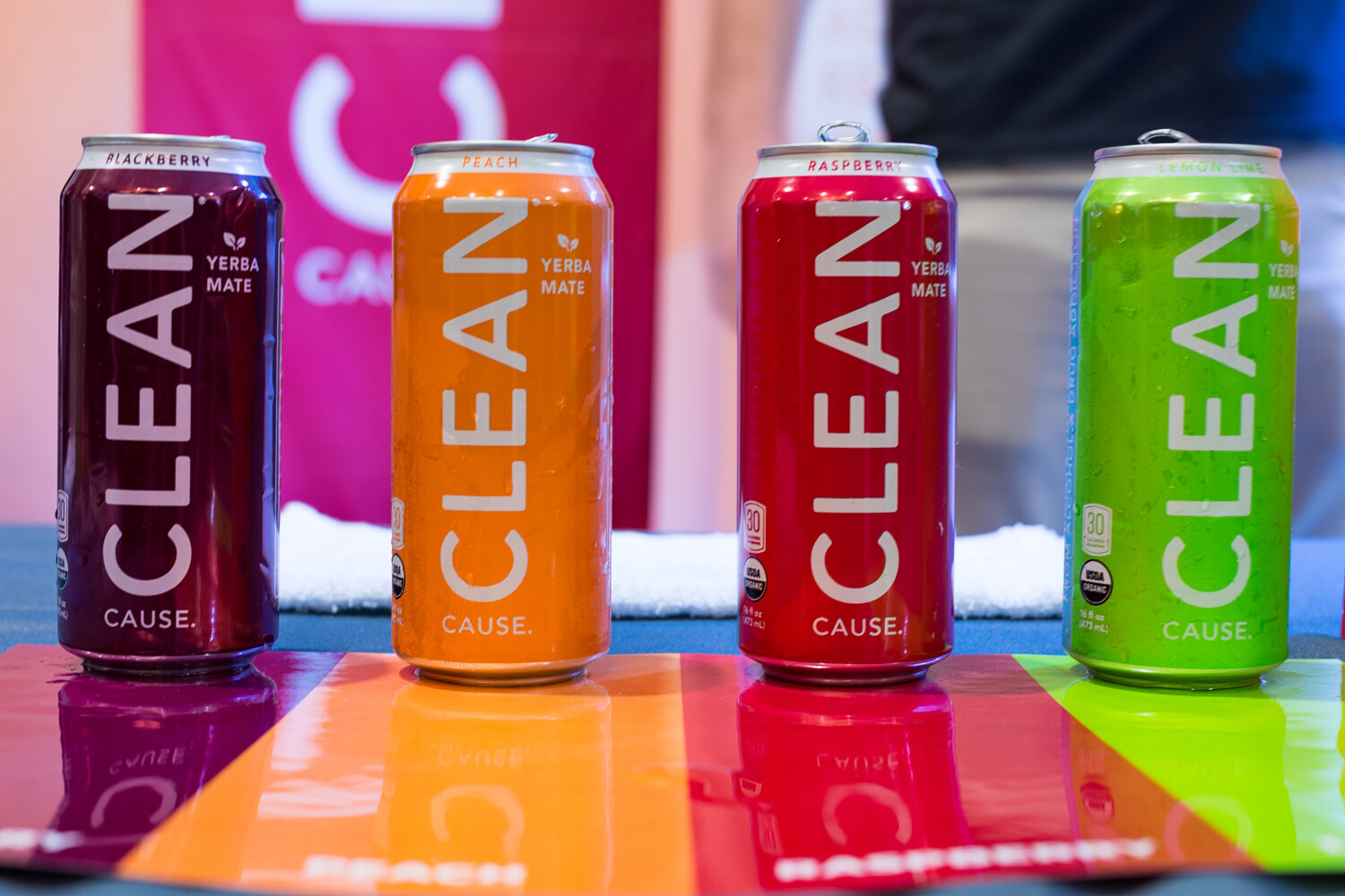 Four tall cans labeled Clean Cause in purple, orange, red and green, standing on a glossy striped table.