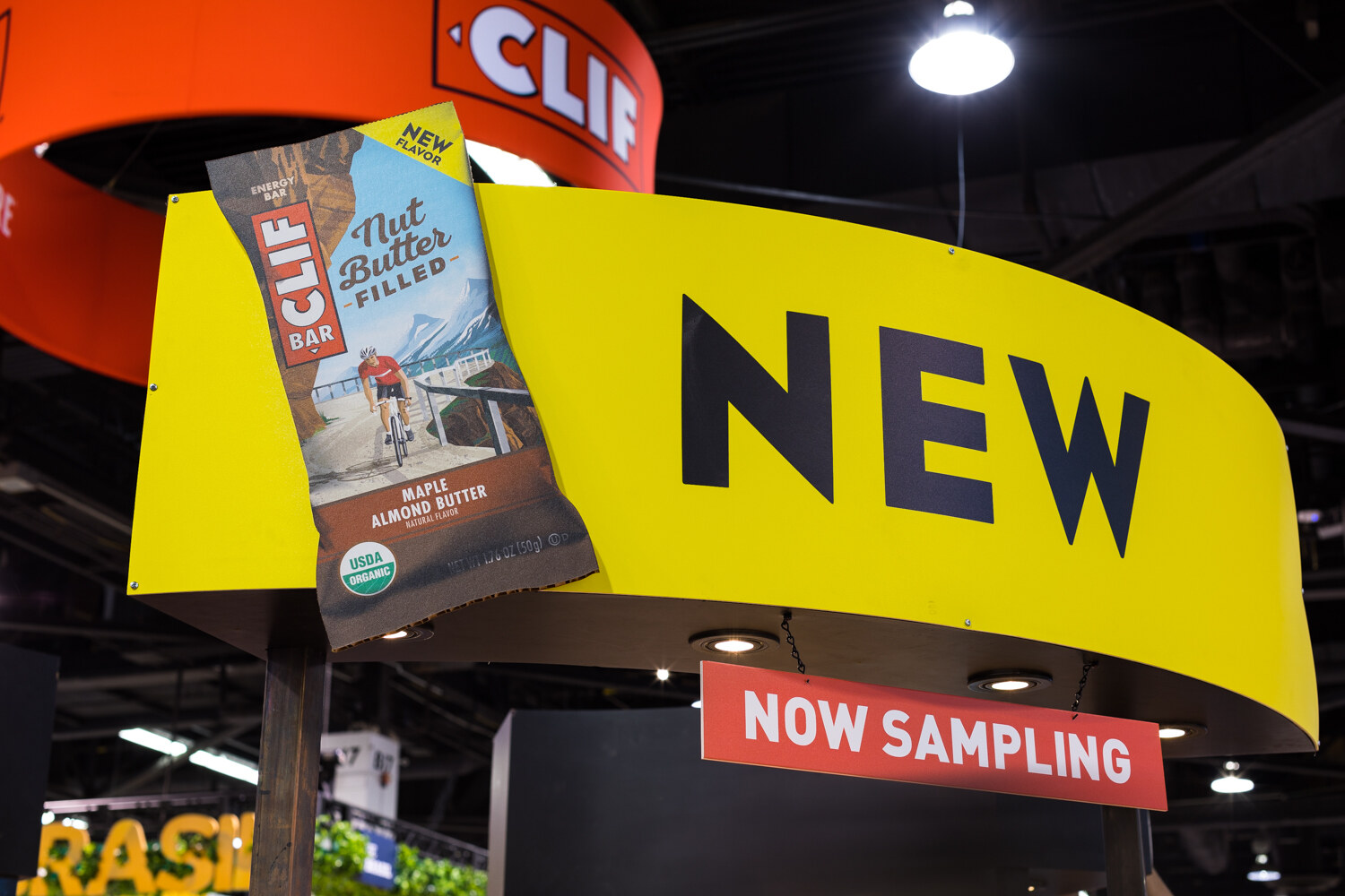 A large yellow curved sign reading NEW with a Clif Bar pinned to it and a red Now Sampling flag, under dark ceiling lights.