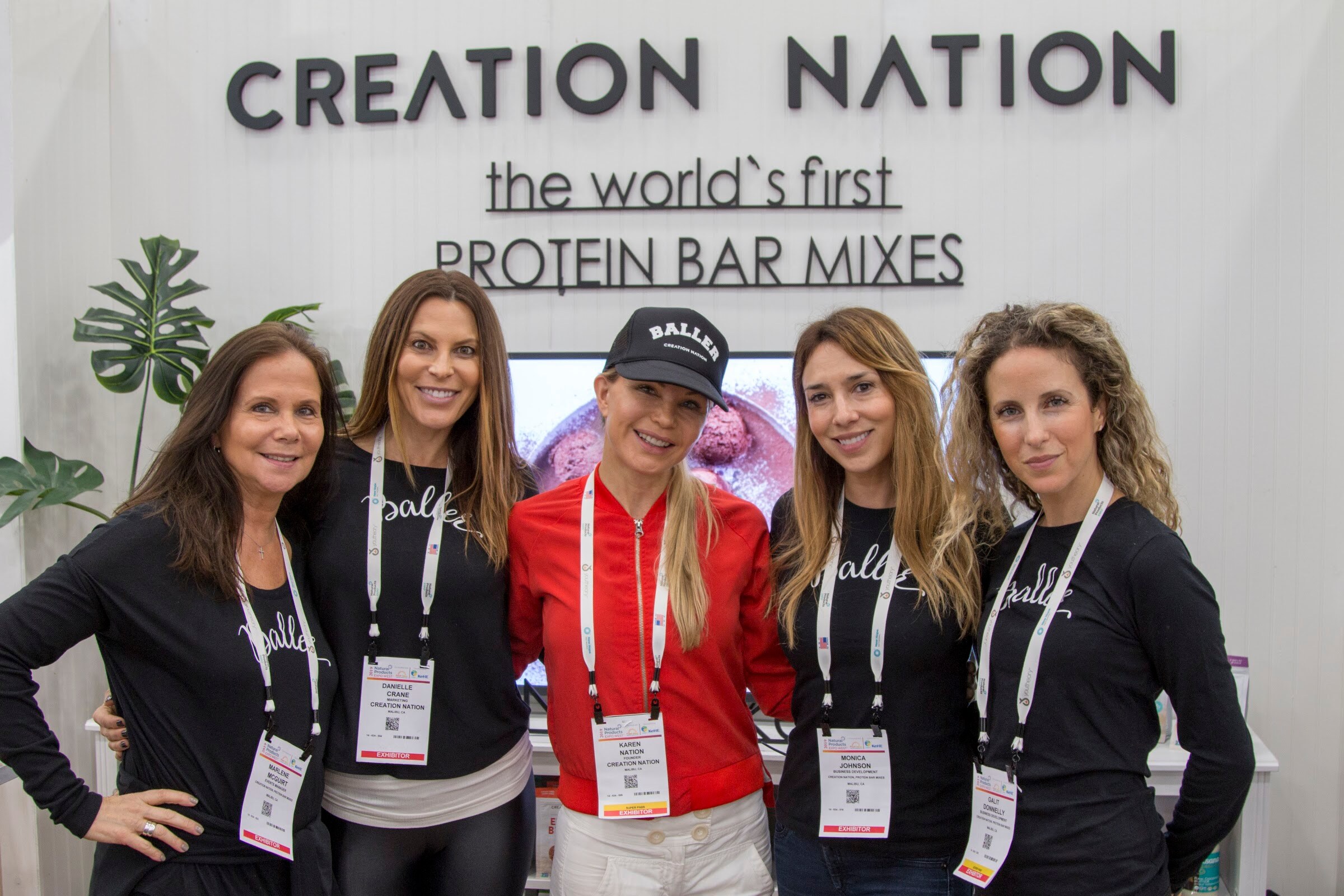 Five women with lanyards and black shirts smiling in front of a white wall with the words Creation Nation and the world's first protein bar mixes.