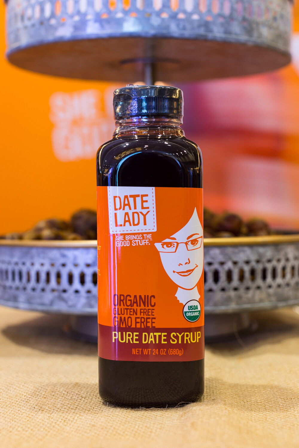 An orange-labeled Date Lady date syrup bottle on a burlap table with an ornate metal stand behind it.