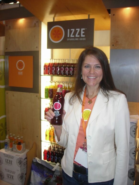 A smiling woman with long brown hair and a white jacket holds a bottle of Izze sparkling juice in front of a lit shelf of bottles and an Izze Sparkling Juice sign.