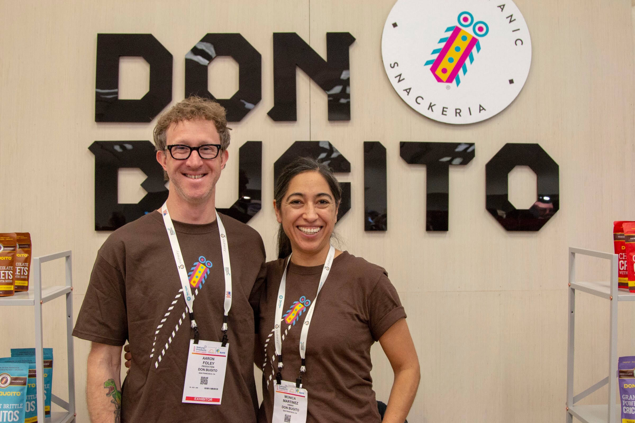 A man with glasses and a woman with long dark hair, both wearing brown T-shirts and lanyards, in front of a Don Bugito sign.