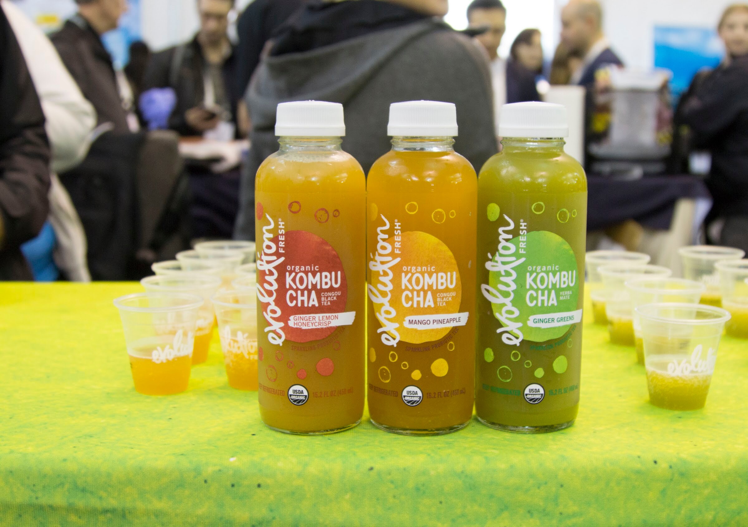 Three kombucha bottles on a lime green tablecloth with small tasting cups and attendees blurred in the background.