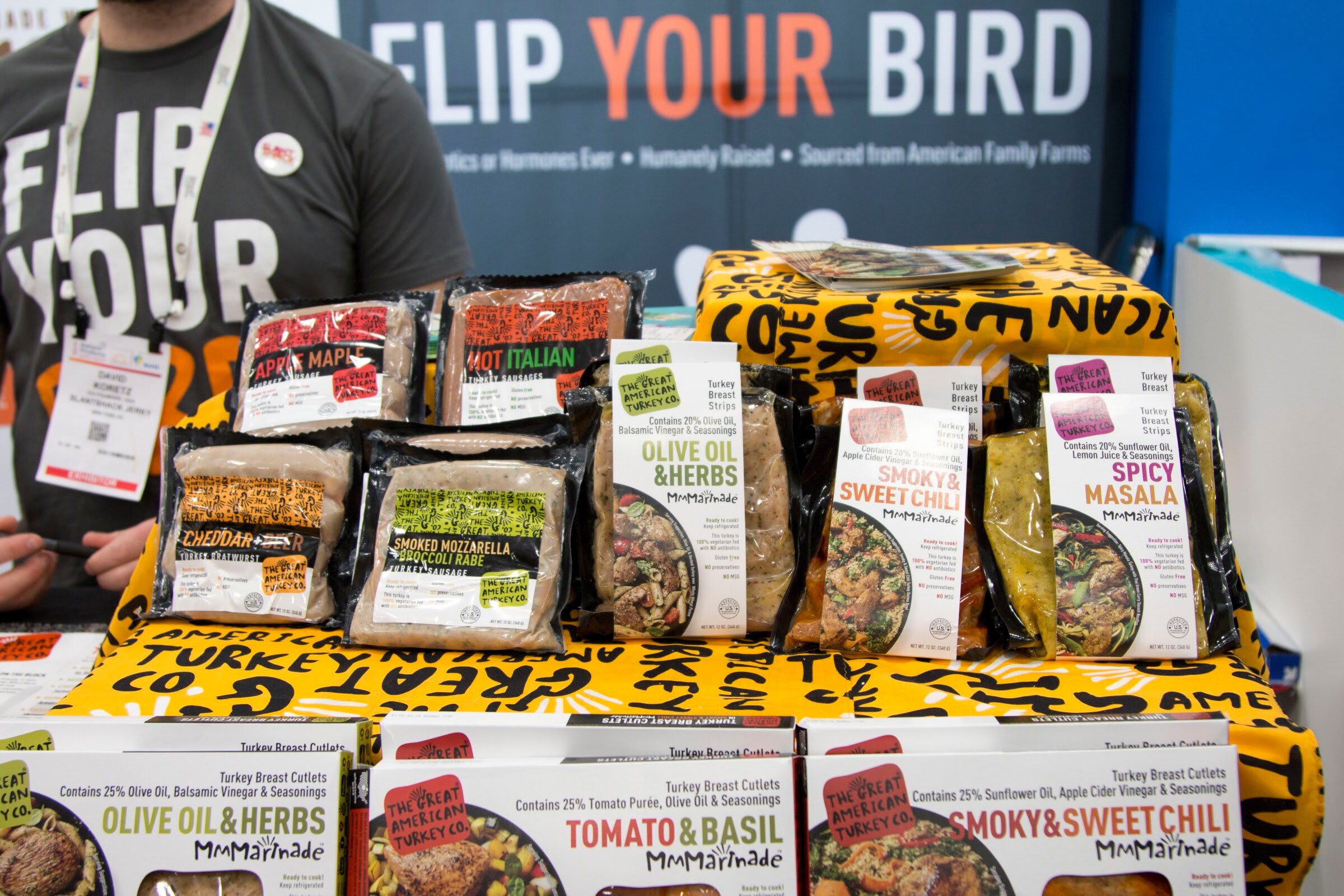 Packages of turkey sausage and marinated cutlets on a black and yellow table with a man in a gray Flip Your Bird T-shirt behind them.