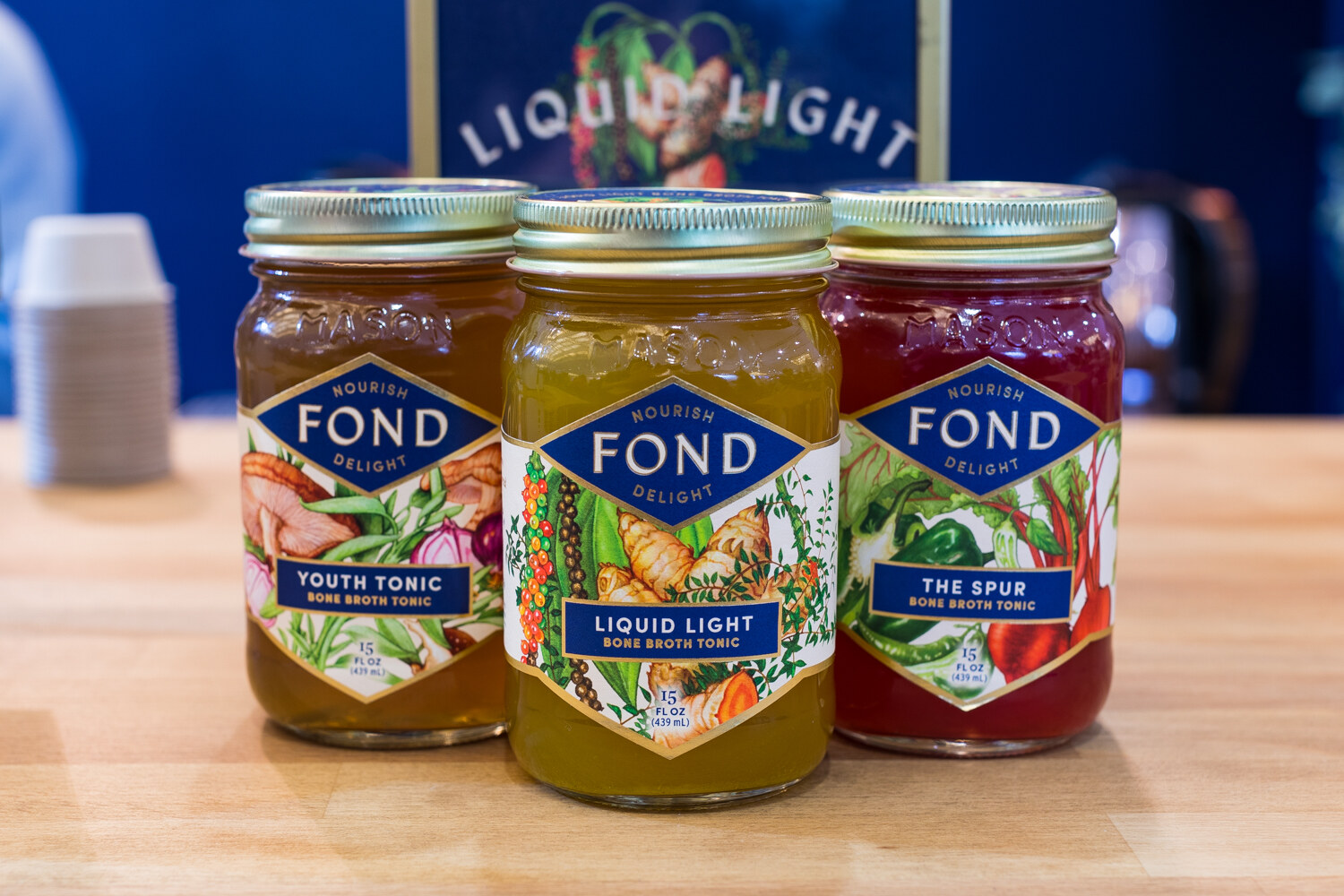 Three jars of Fond bone broth tonic with diamond labels on a butcher block counter in front of a Liquid Light sign.