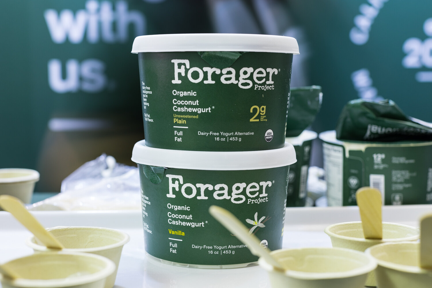 Two stacked green tubs of Forager Project Organic Coconut Cashewgurt with beige sample bowls and spoons in front of a green banner.