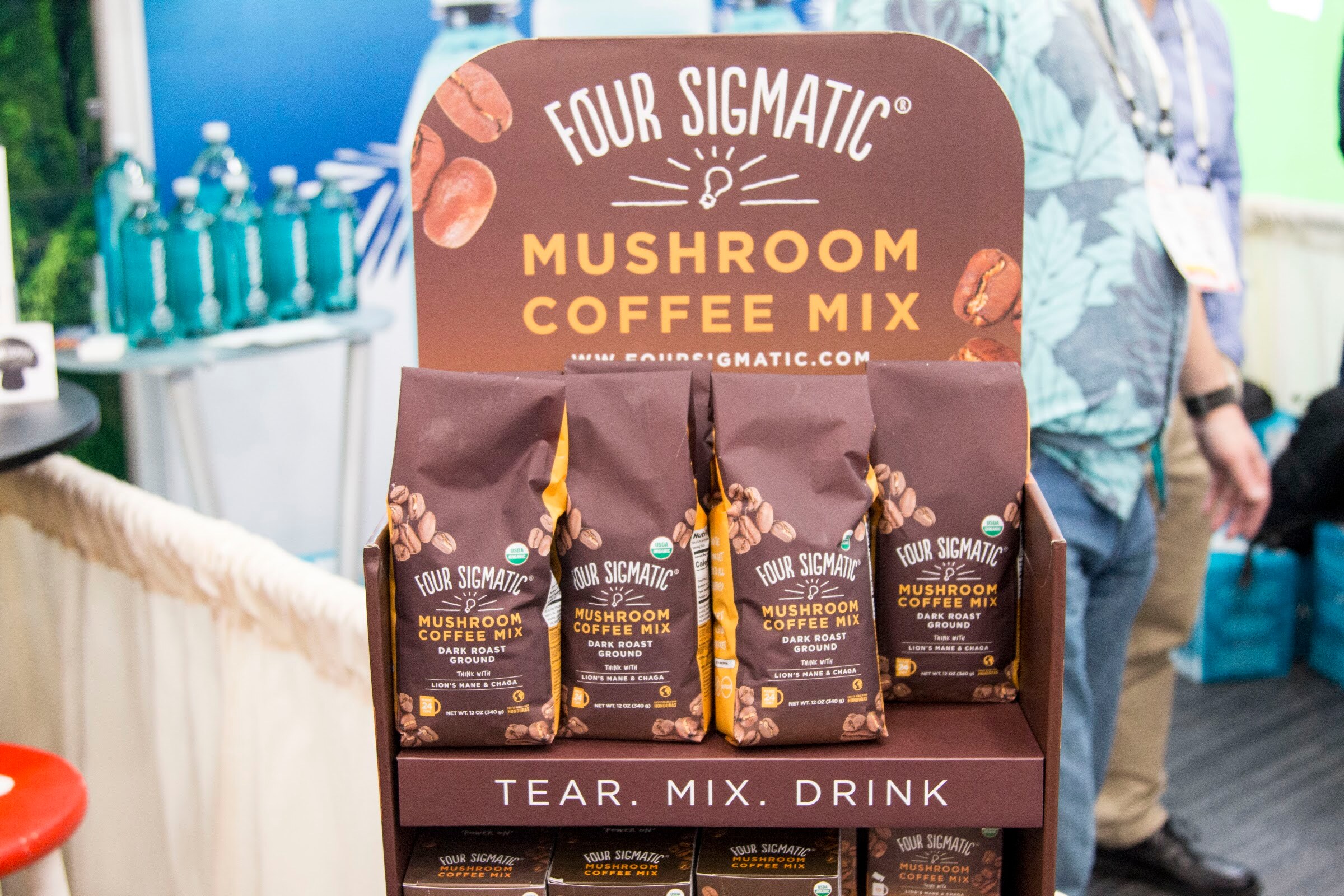 Four brown bags of Four Sigmatic mushroom coffee mix in a tear mix drink display with the lower body of a man in a patterned shirt behind.