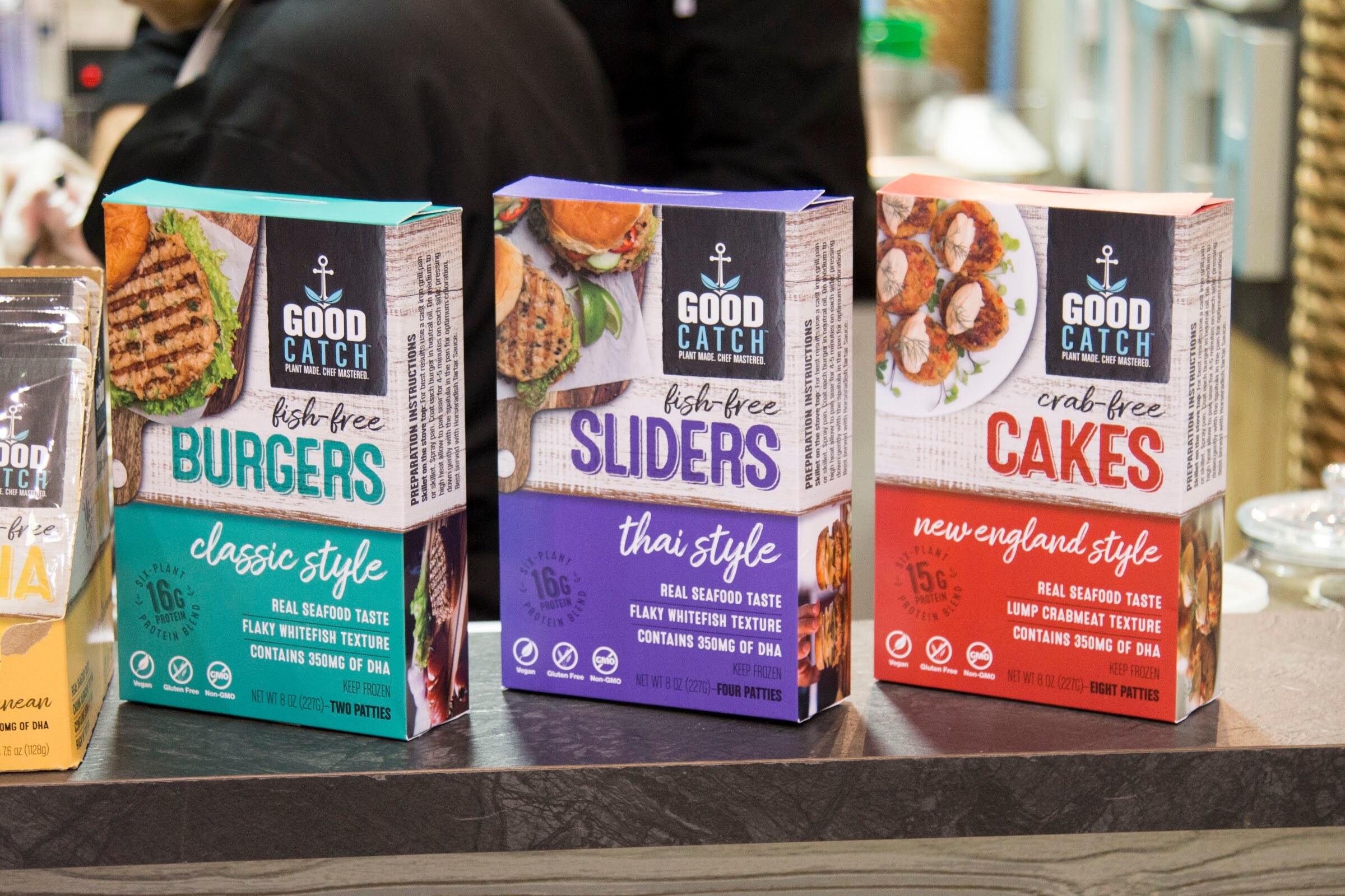 Three boxed products, Good Catch Burgers in teal, Sliders in purple and Cakes in red, with a person in dark clothing blurred behind.