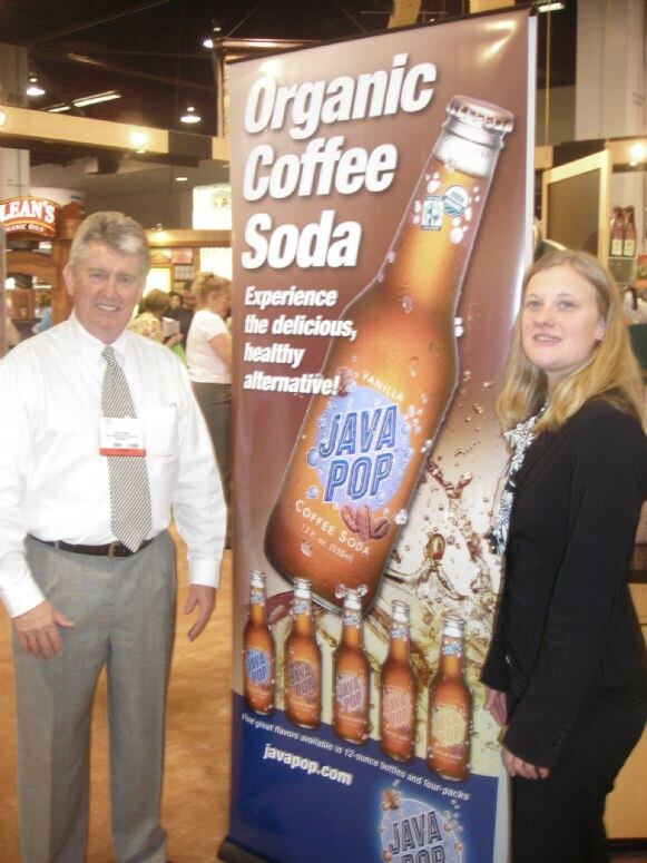 A man in a white shirt and striped tie and a woman with long blonde hair in a black jacket stand on either side of a Java Pop Organic Coffee Soda banner showing bottles.