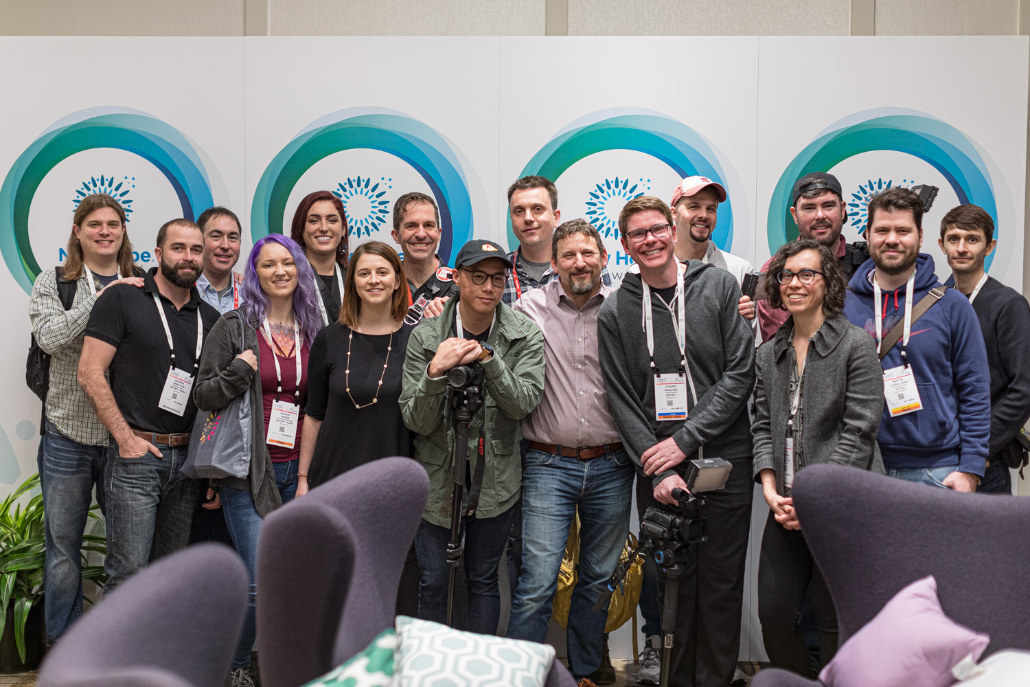 A crowd of smiling people standing and kneeling in front of a white wall with teal circle graphics, with purple chairs in the foreground.