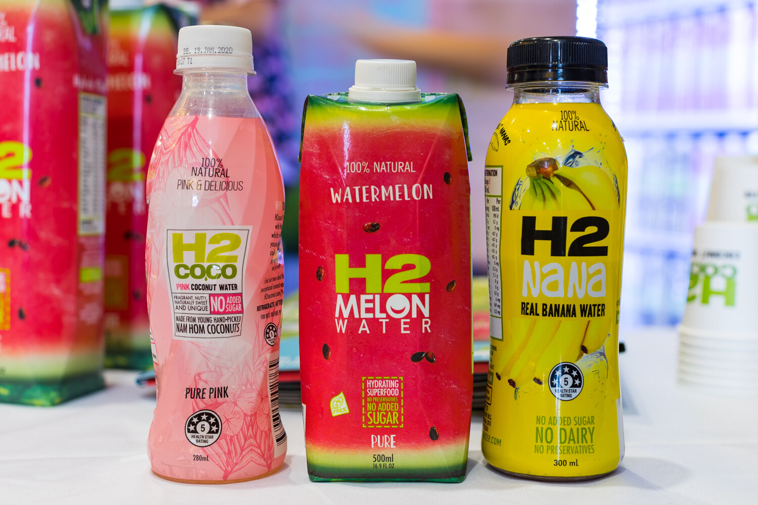 Three H2 drinks, a pink bottle, a watermelon-patterned carton and a yellow bottle, on a white table with cups behind.