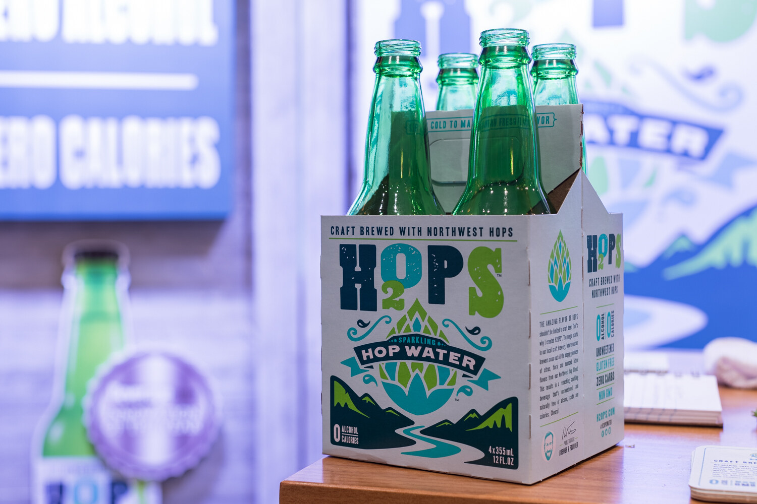 A cardboard carrier of four green bottles labeled H2Ops sparkling hop water on a wooden counter with a blurred blue sign behind.