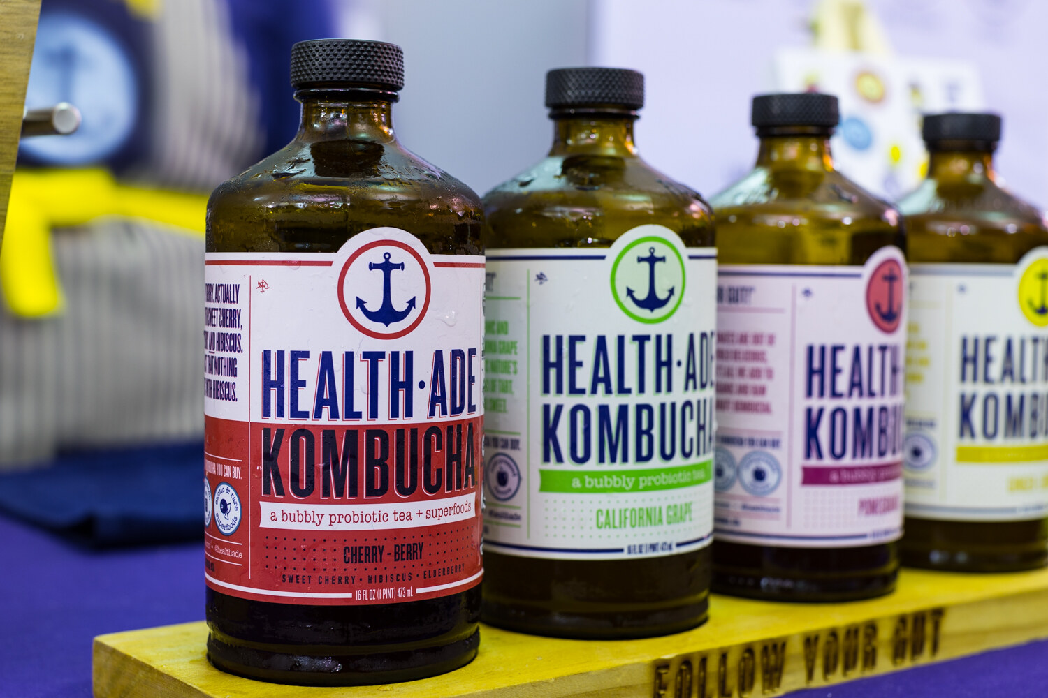 Four amber Health-Ade Kombucha bottles with colorful labels on a yellow wooden riser with the words Follow Your Gut.