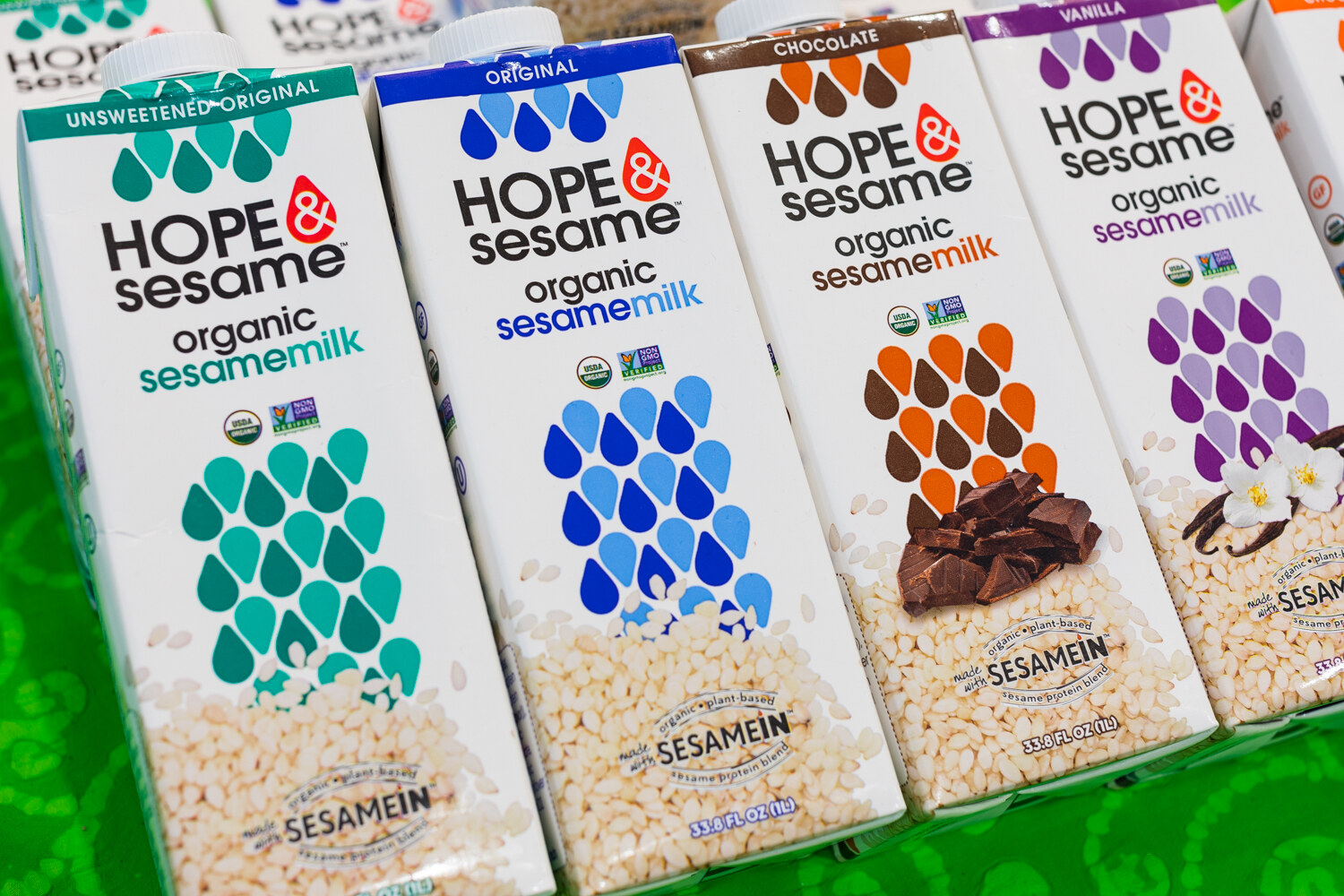 Four angled Hope and Sesame organic sesamemilk cartons in teal, blue, brown and purple on a green surface.