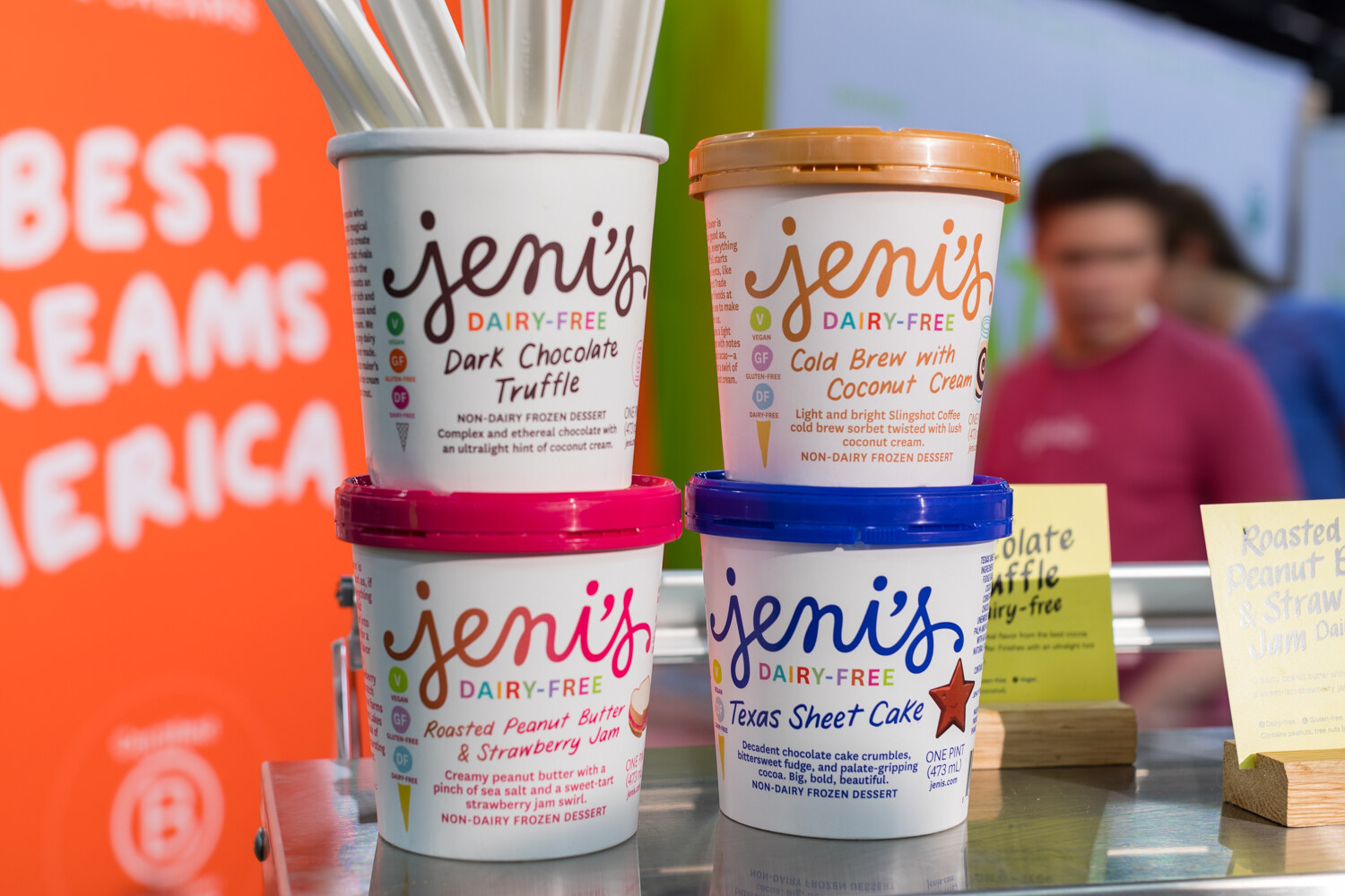 Four Jeni's ice cream tubs in white, orange, pink and blue lids stacked on a metal counter with an orange wall behind.