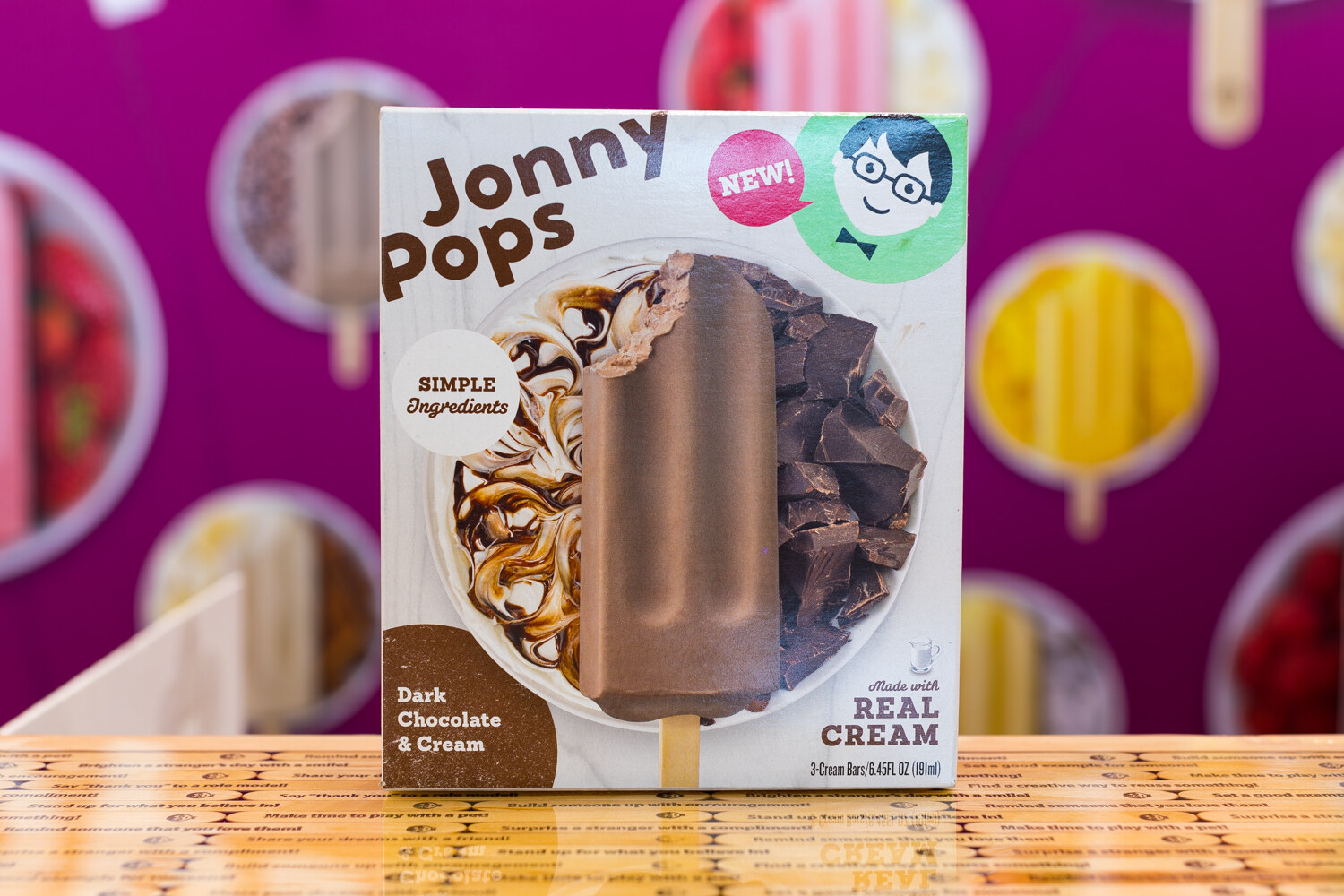 A JonnyPops box with a chocolate bar photo on a table in front of a magenta backdrop of pops.