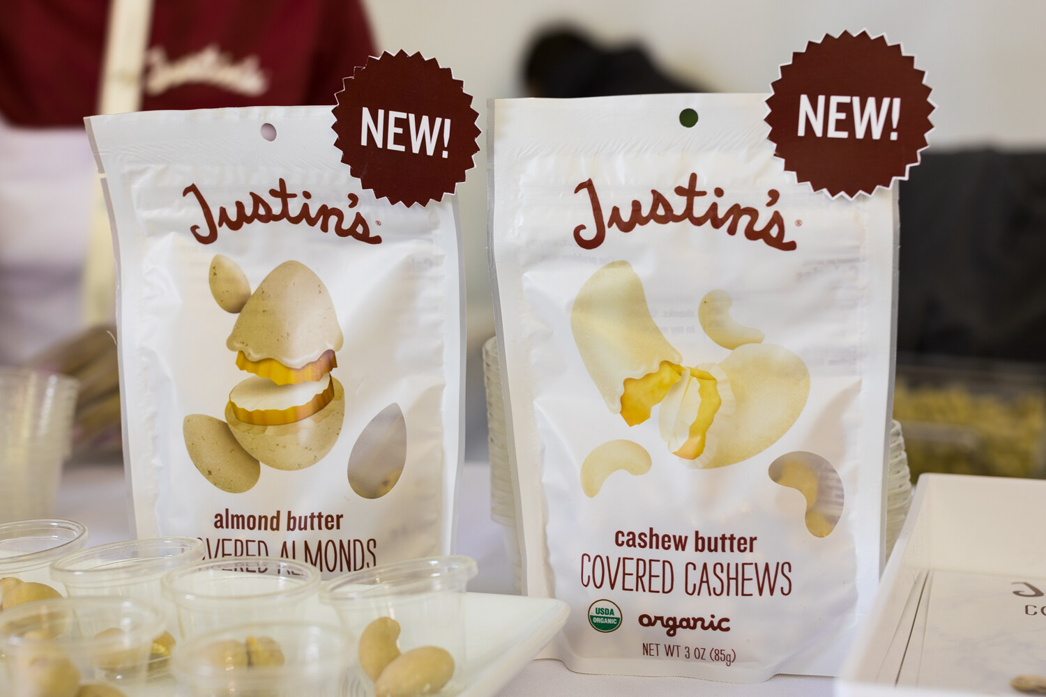 Two white Justin's pouches with red NEW starbursts, beside small clear sample cups holding nuts.