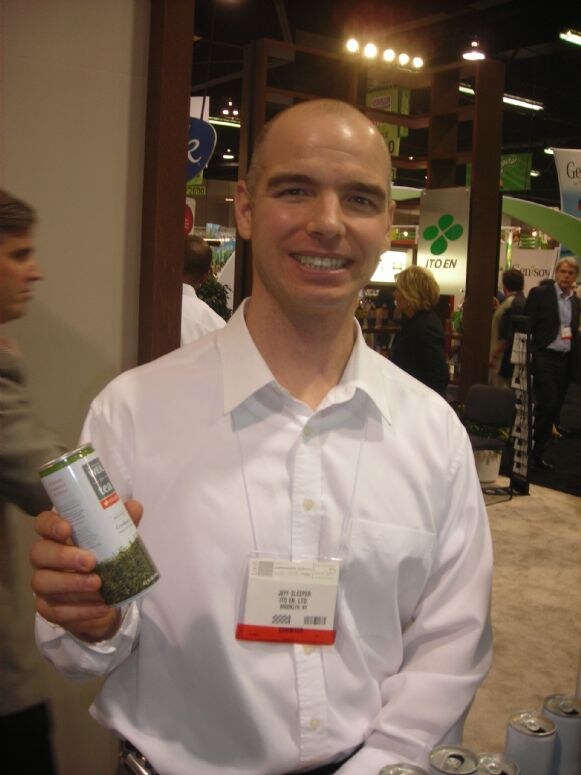 Jeff Sleeper of Ito En, Ltd., a smiling bald man in a white shirt with a badge, holds a canned tea drink at the Ito En booth with its green clover logo behind him.