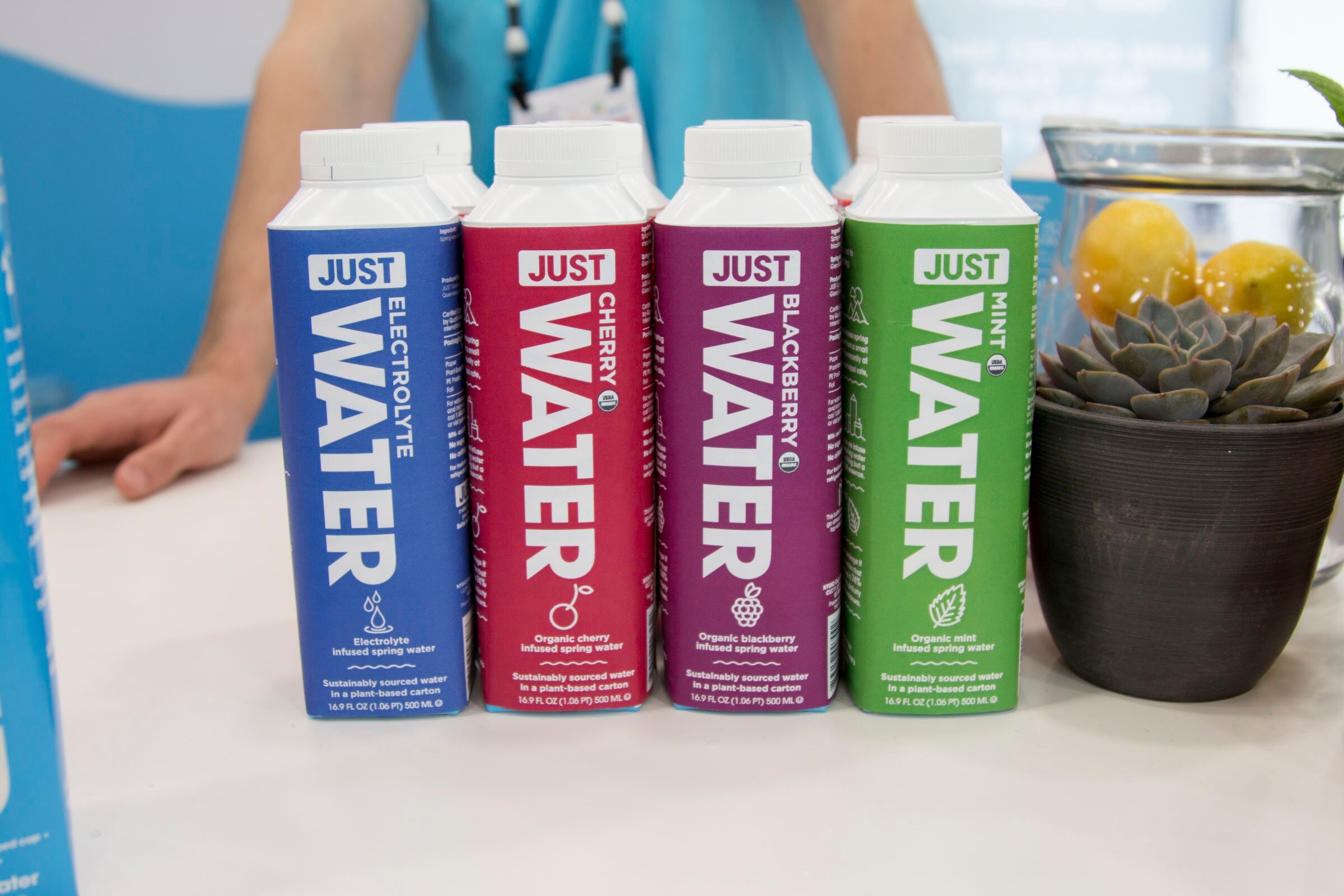 Four boxed water cartons in blue, red, purple and green with a succulent and lemons in a jar to the side.
