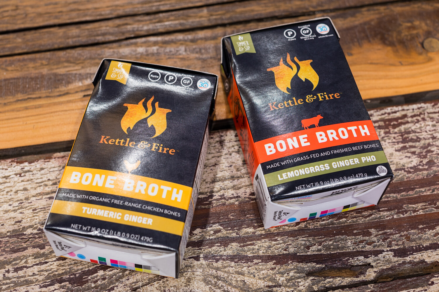 Two black Kettle and Fire bone broth cartons with yellow and orange labels lying on rough wood.