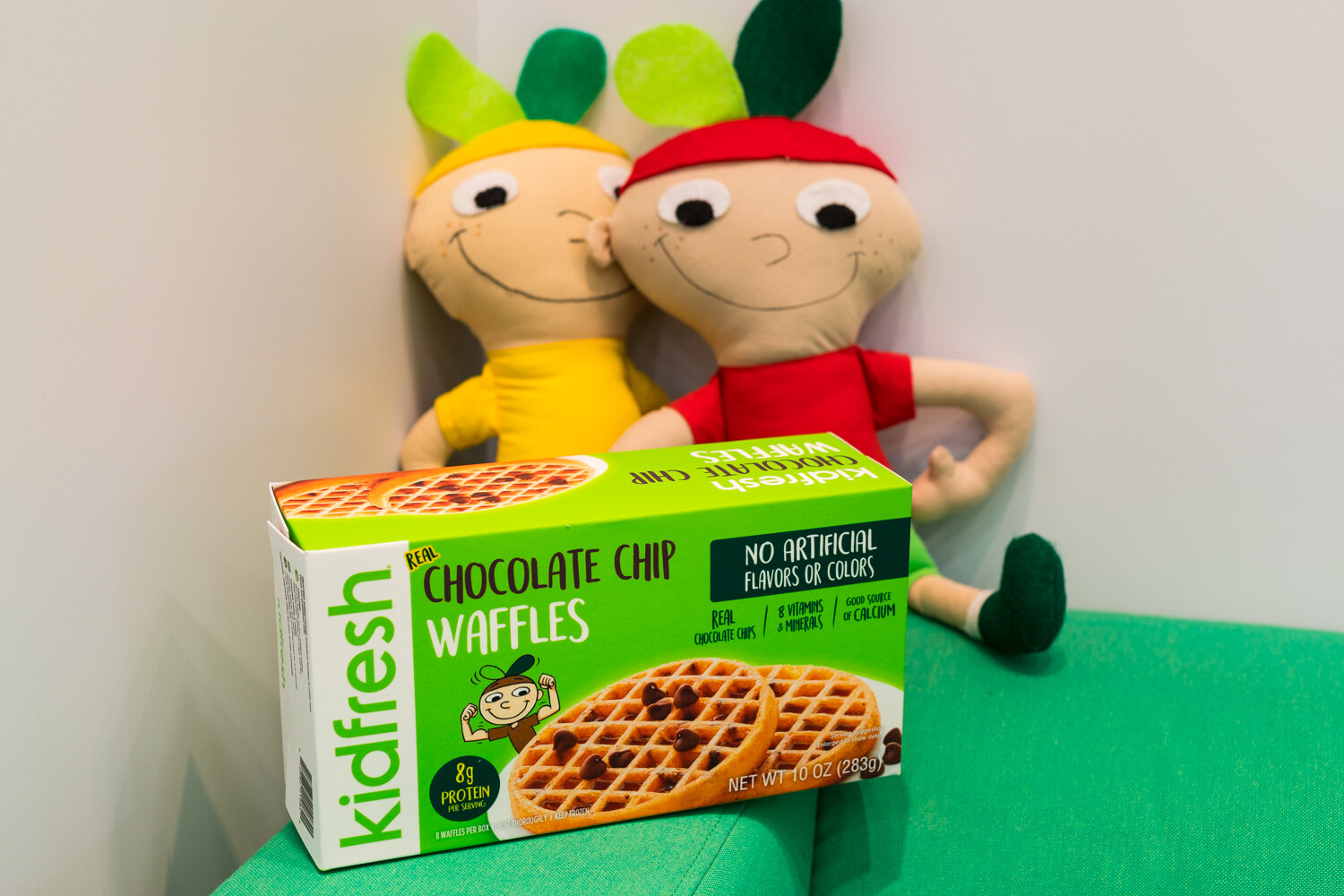 A green Kidfresh waffles box leaning on a green cushion with two stuffed doll characters behind it.