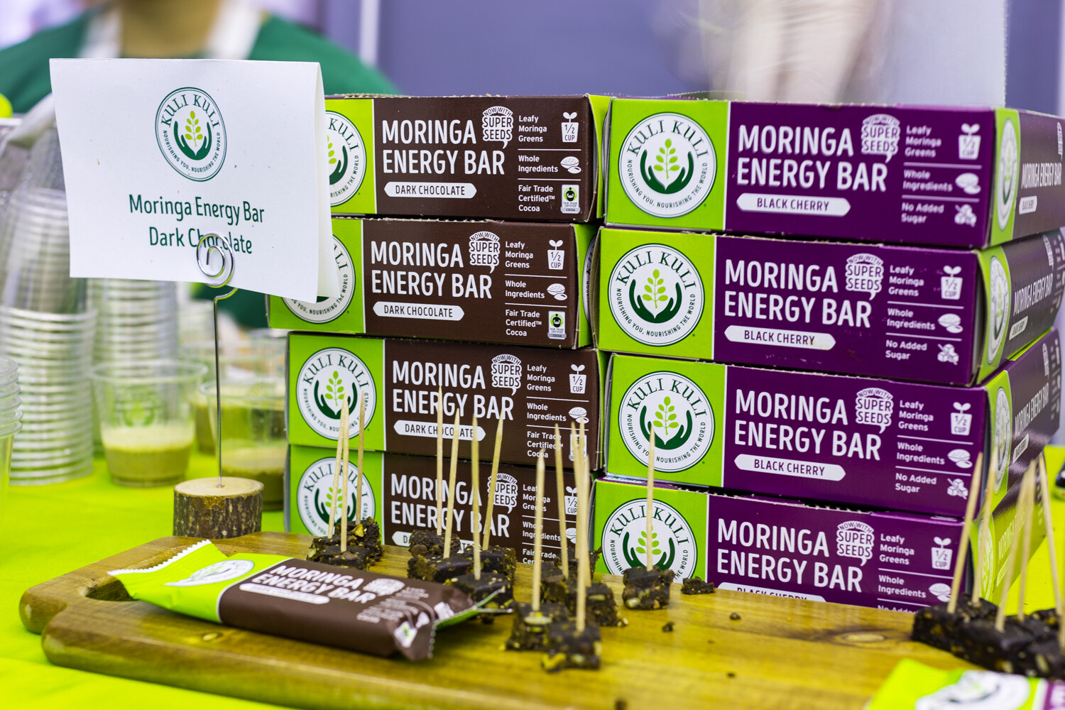 Stacks of green and purple Kuli Kuli Moringa Energy Bar boxes with a table sign and wooden board of samples on skewers.