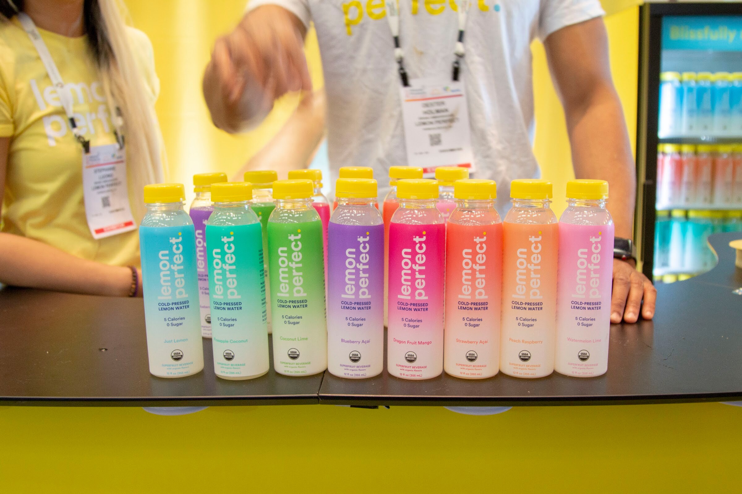 A row of colorful Lemon Perfect bottles with yellow caps on a black counter with two staffers in yellow in the background.