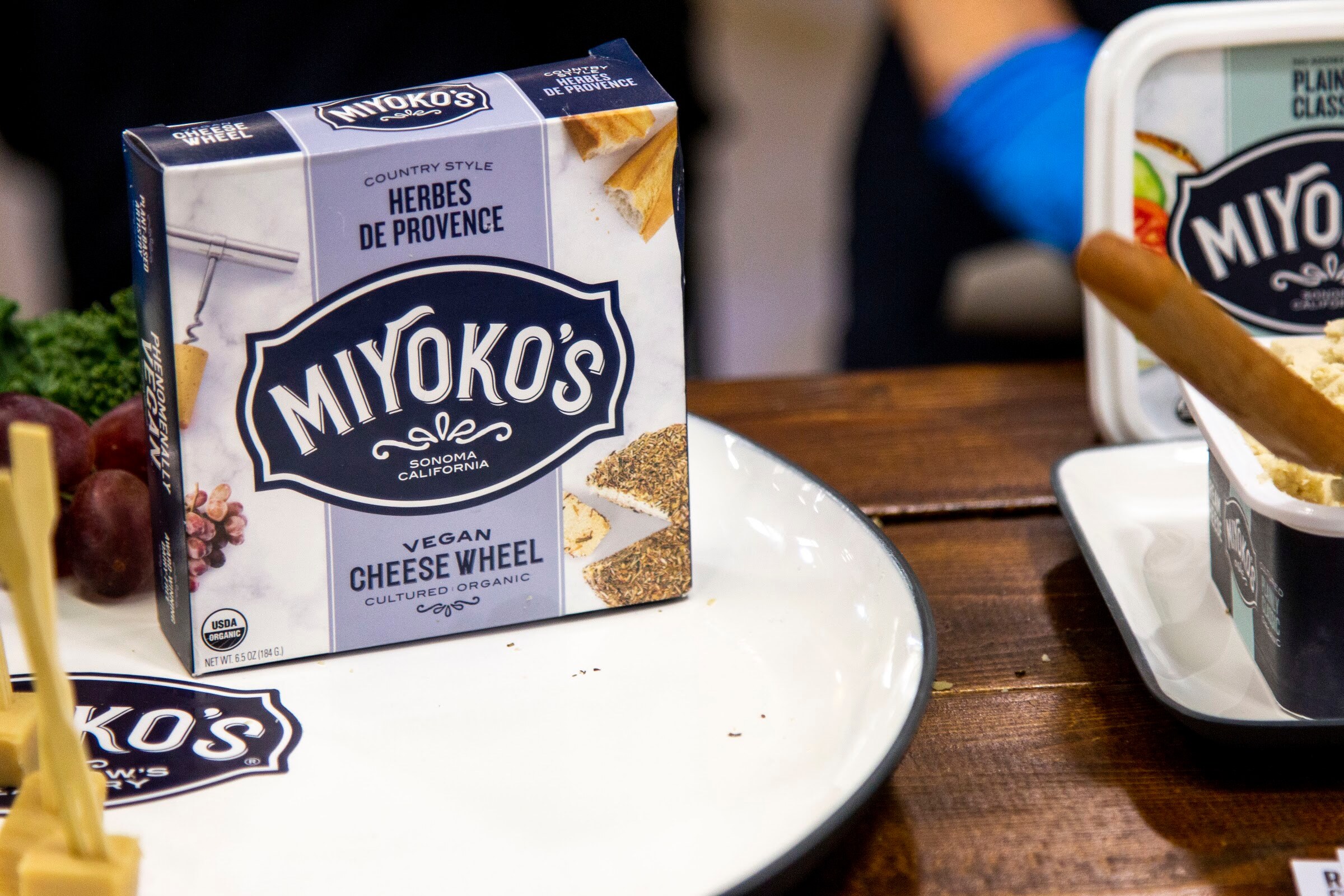 A Miyoko's cheese wheel box on a white plate with sample picks, with a gloved hand blurred in the back.