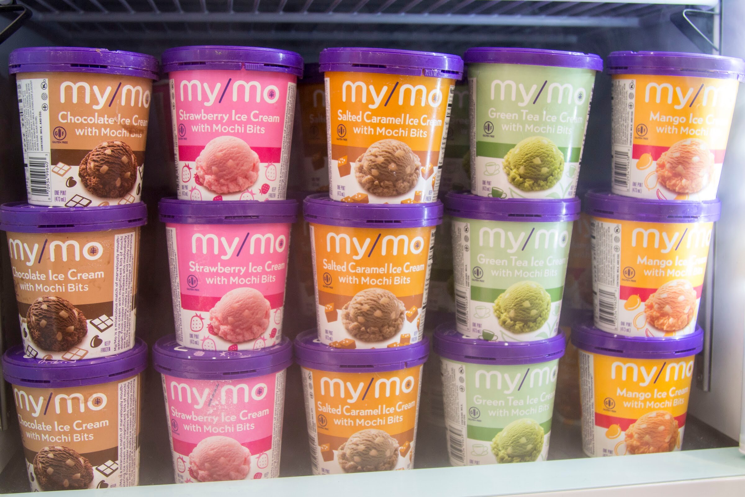 Stacked pints with purple lids and flavor-colored labels in three rows inside a glass freezer.