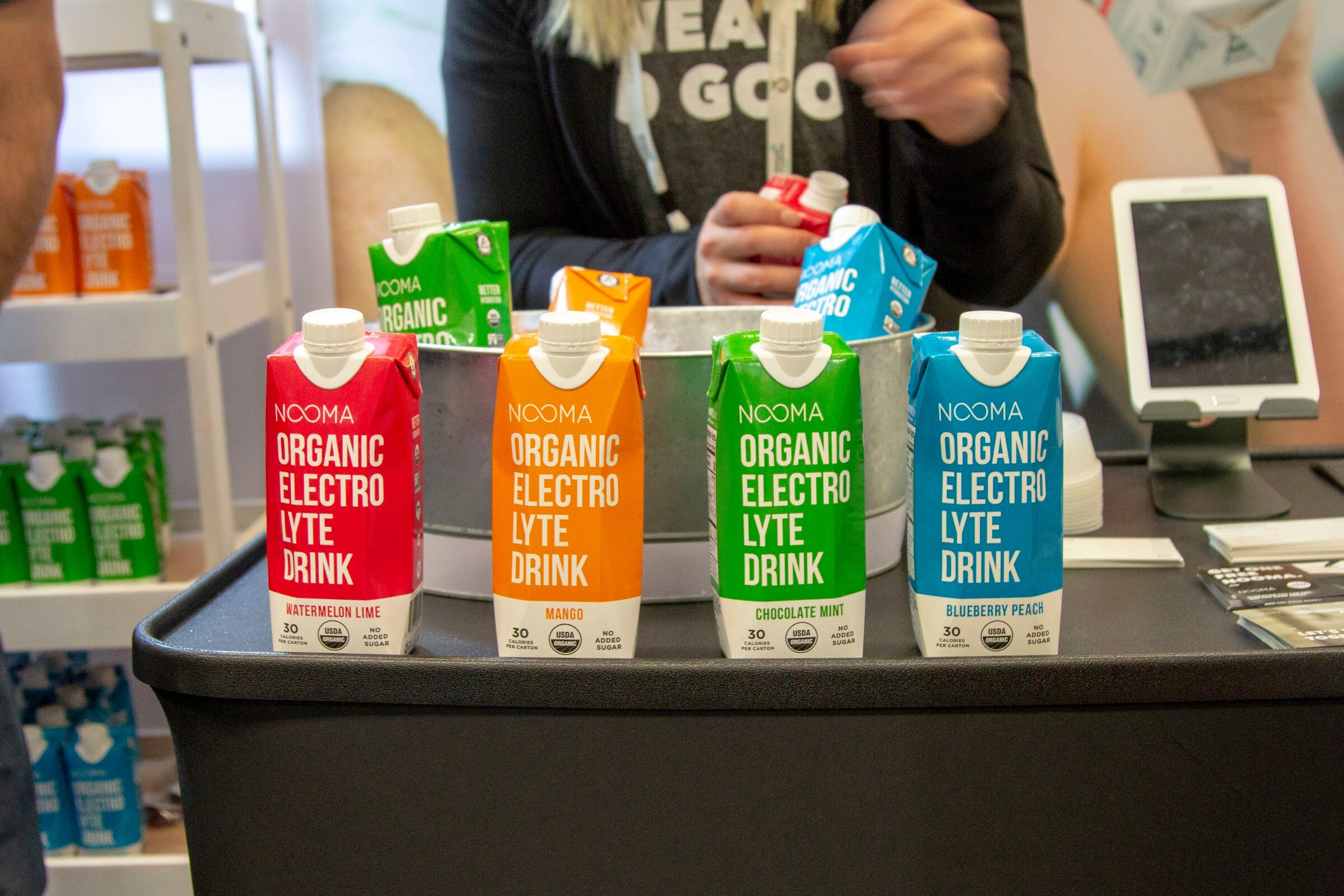 Four Nooma cartons in red, orange, green and blue with another staffer's arms and a tablet behind them.