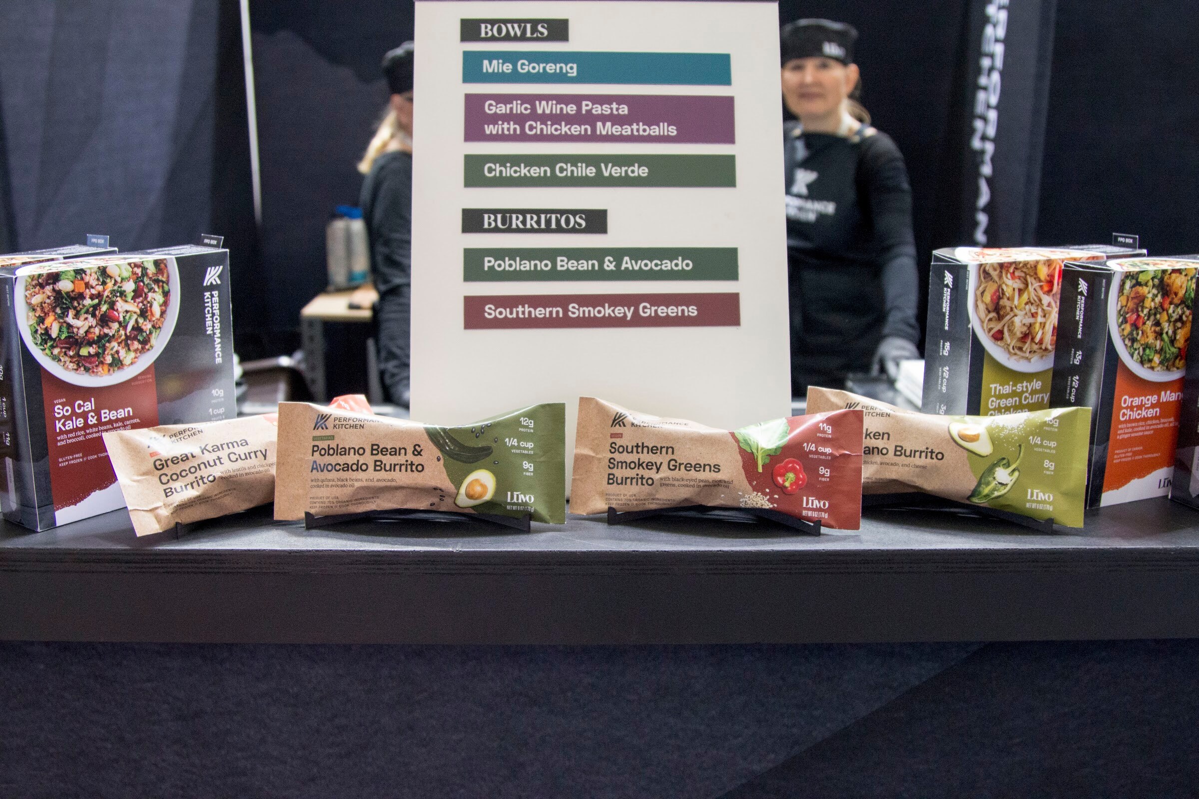 Wrapped burritos and boxed bowls on a black table in front of a menu board, with a cook in a black cap visible behind.