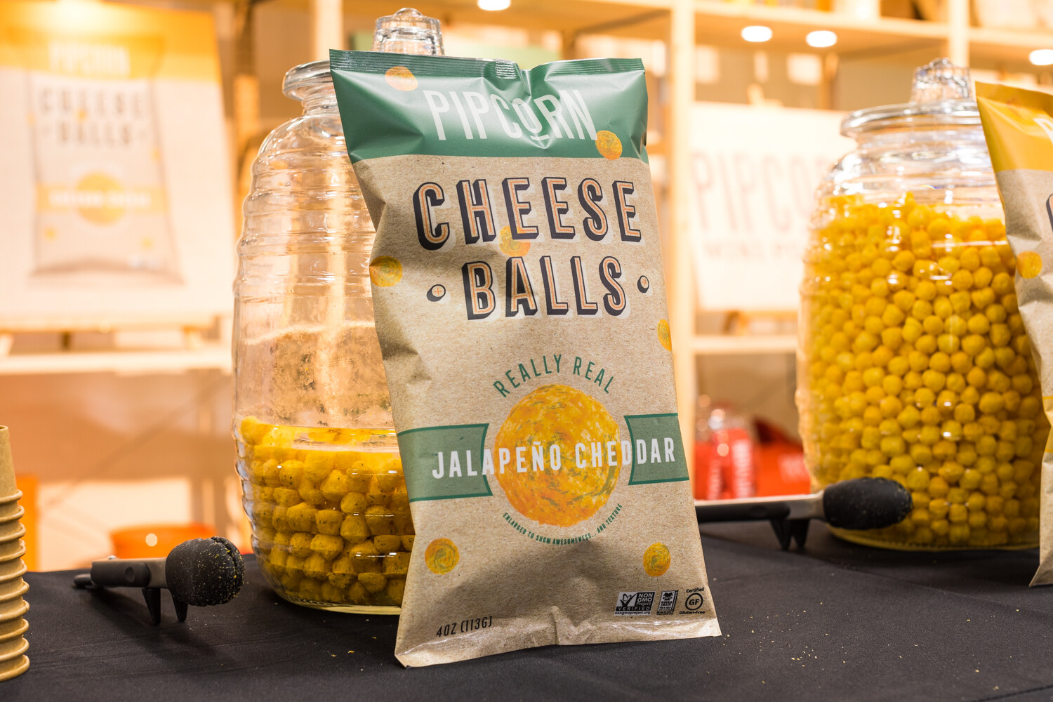 A kraft Pipcorn bag leaning on a glass jar of yellow puffs, with a second jar and sample tongs on a black table.