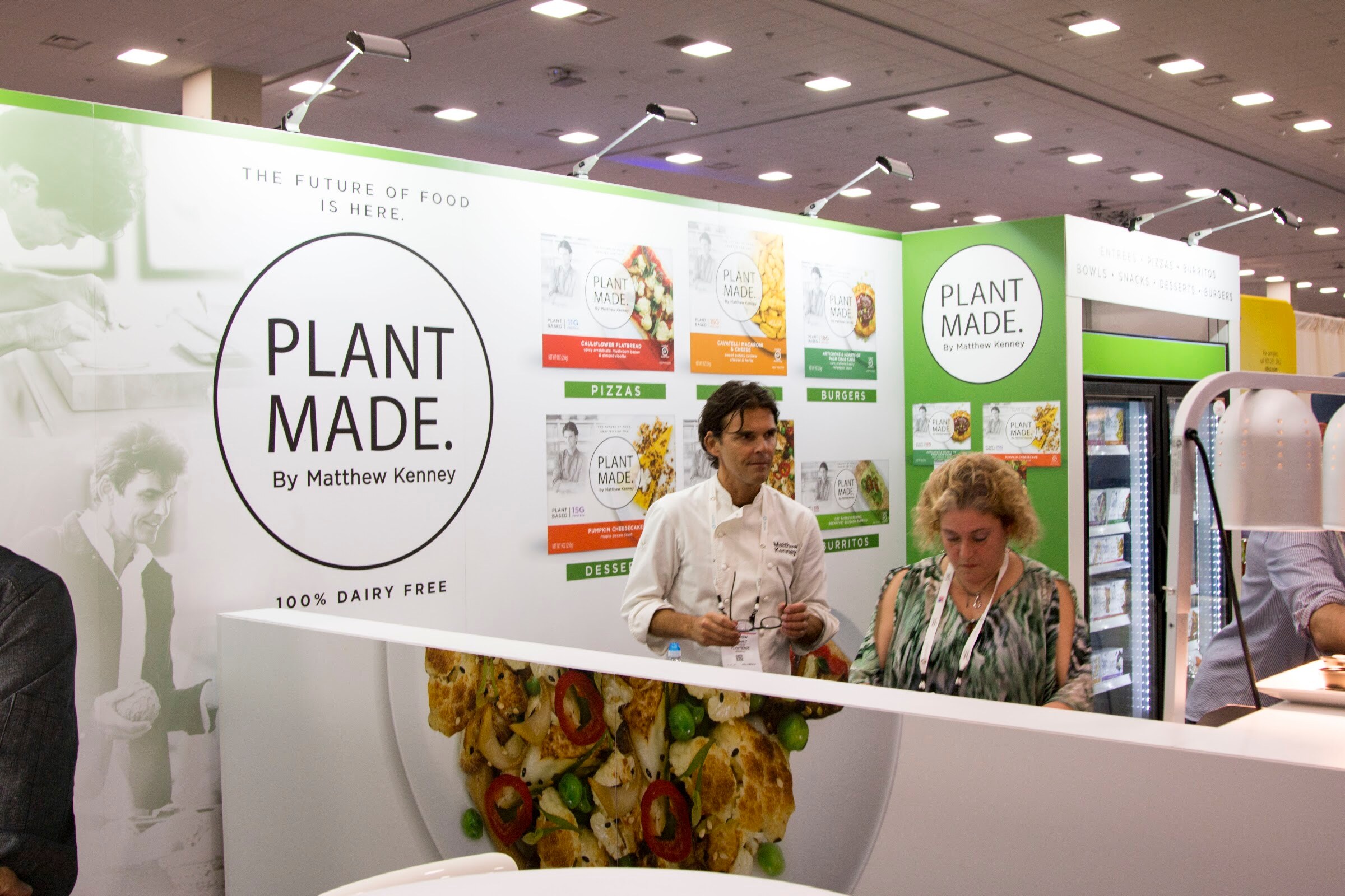 A white booth with a large Plant Made sign and food photos, a chef in a white jacket and a woman with a lanyard at the counter, under the convention hall ceiling lights.
