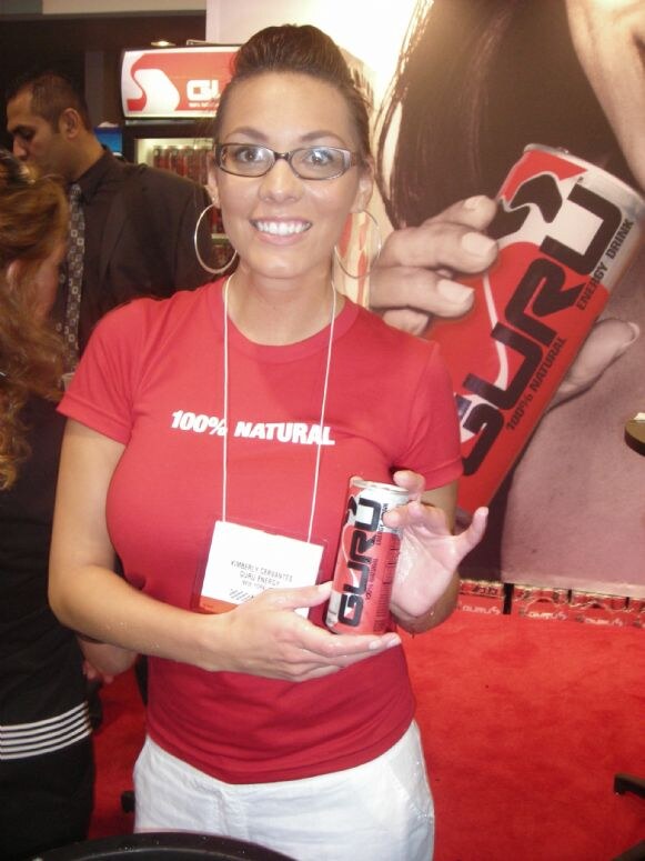 Kimberly Cervantes of Guru Energy smiles in a red "100% Natural" t-shirt and glasses, holding a Guru energy drink can in front of a large Guru can banner on red carpet.