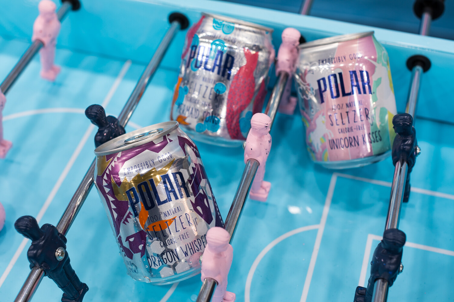 Three Polar seltzer cans sitting on a blue foosball table among pink and navy players and rods.