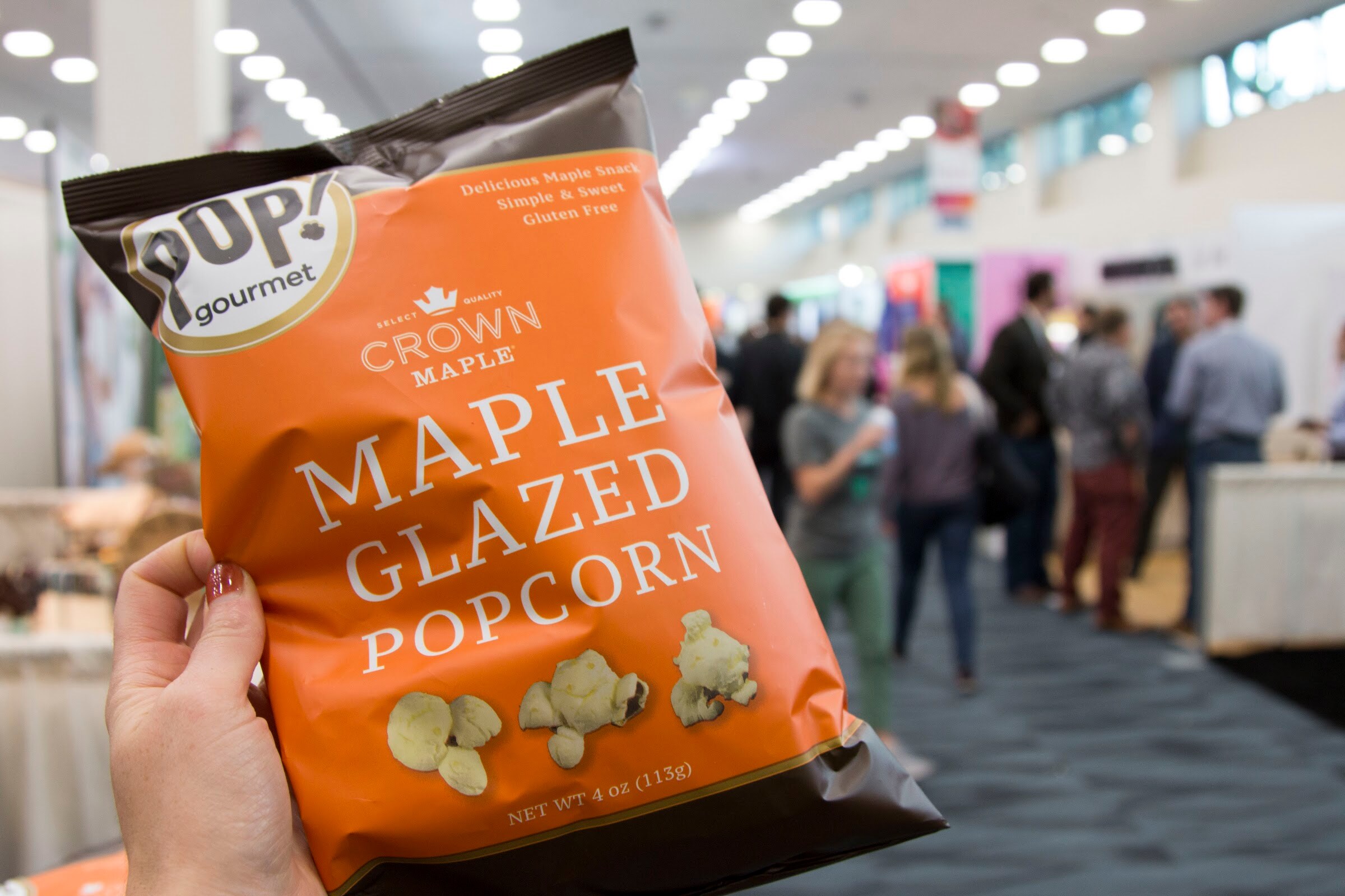 An orange bag of Pop Gourmet Crown Maple glazed popcorn held in a hand with a blurred crowded hall behind.
