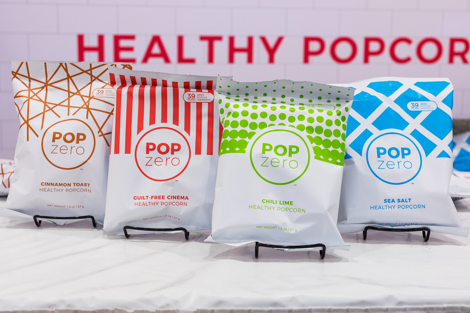 Four white Pop Zero popcorn bags with colored patterns on black wire stands in front of a white tiled wall.