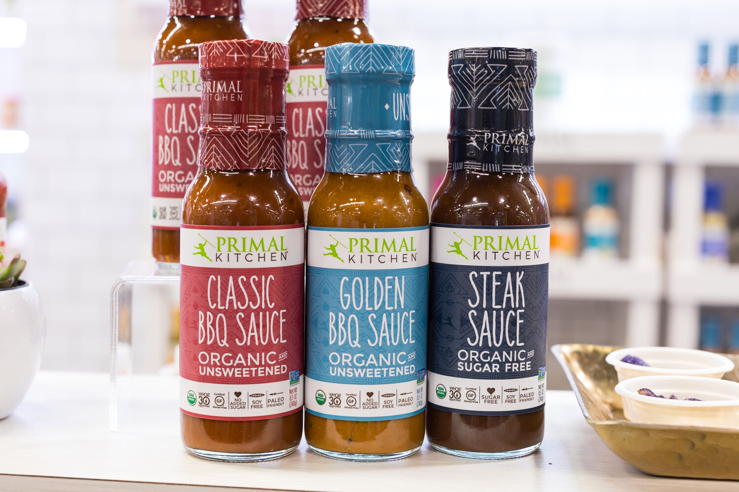 Three bottles of Primal Kitchen sauce with red, blue and navy labels on a white counter with a gold tray of cups.
