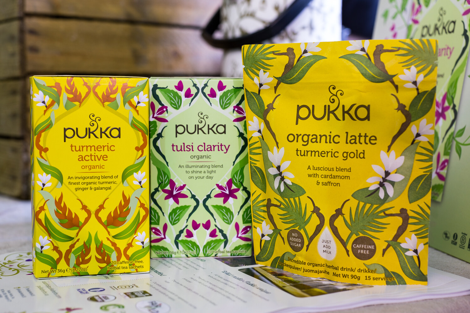 Yellow and green Pukka tea boxes and a larger yellow pouch of organic latte turmeric gold on a table in front of a wooden crate.