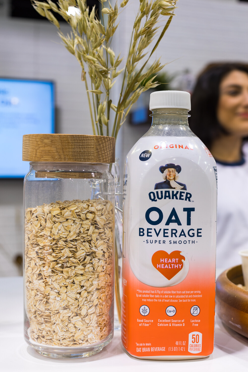 A Quaker Oat Beverage bottle with an orange label next to a glass jar of oats, with oat stalks above and a smiling woman blurred behind.