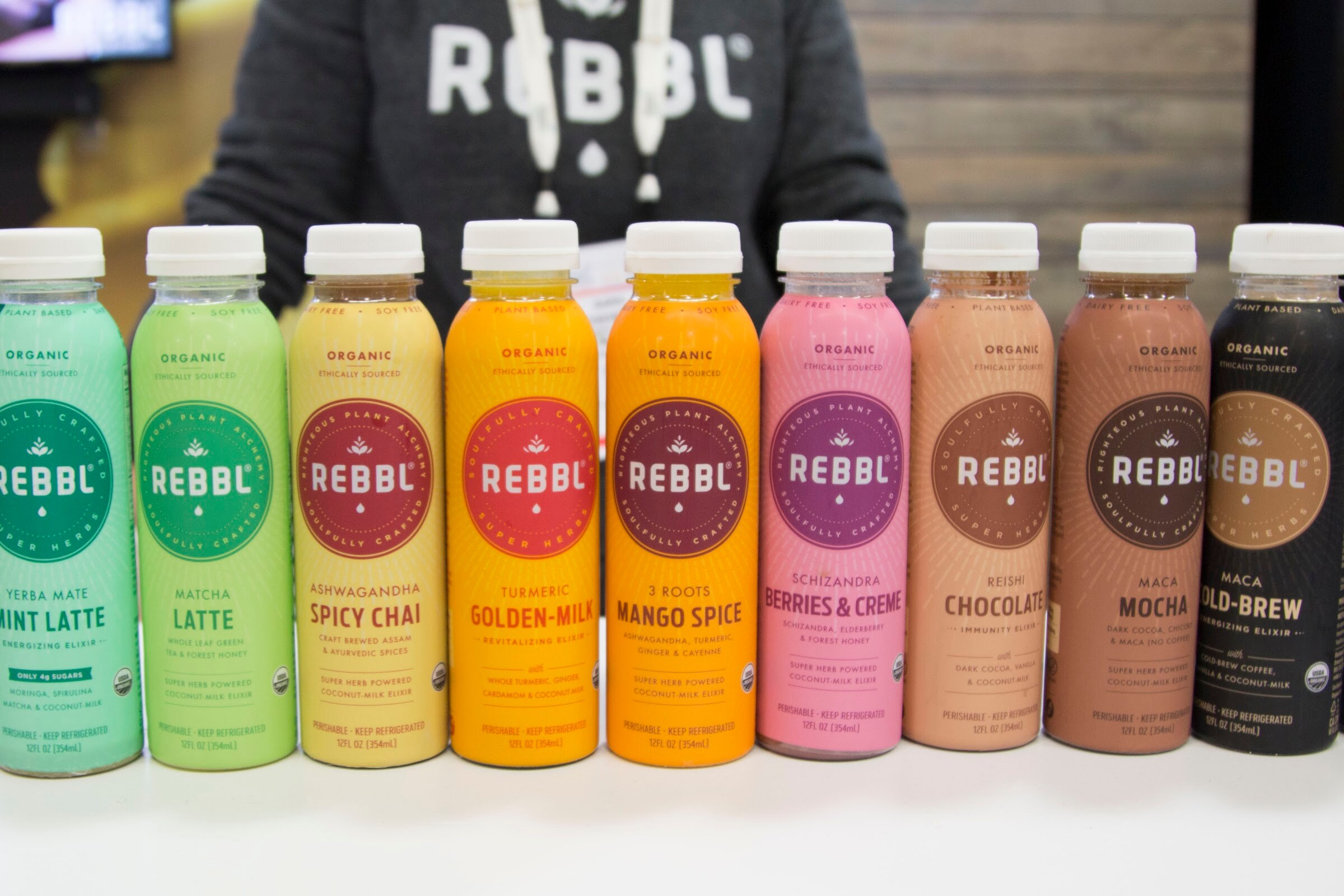 Nine colorful REBBL bottles in a row on a white counter with a staffer in a REBBL sweatshirt behind.