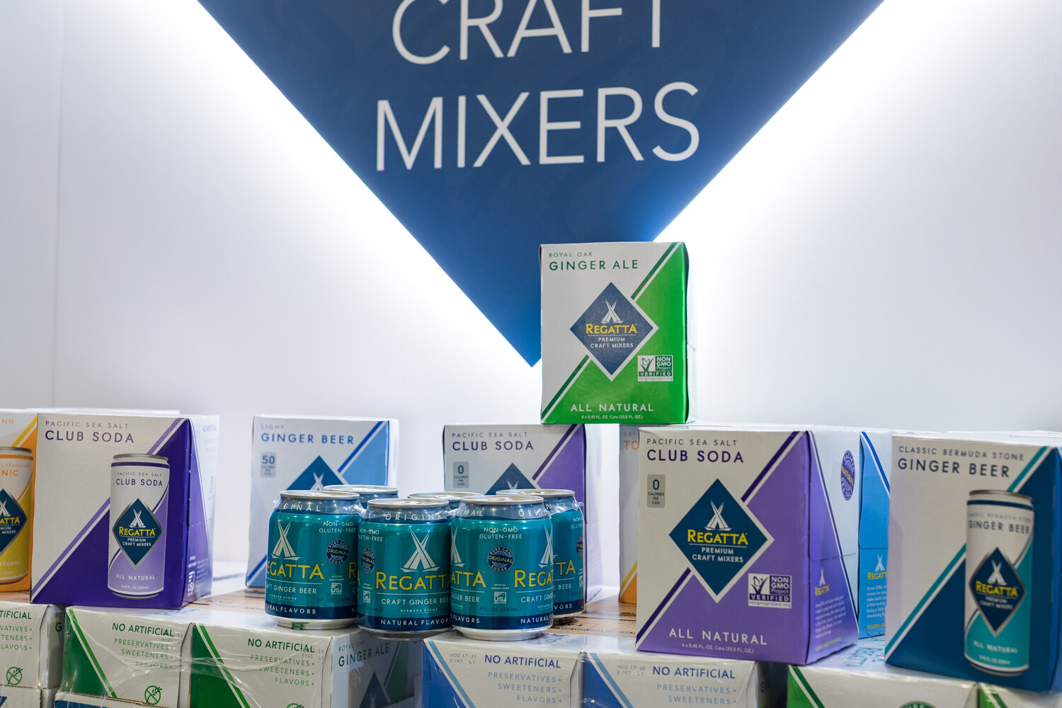 Boxes and cans of Regatta craft mixers stacked on shelves beneath a white wall with a blue triangular Craft Mixers sign.