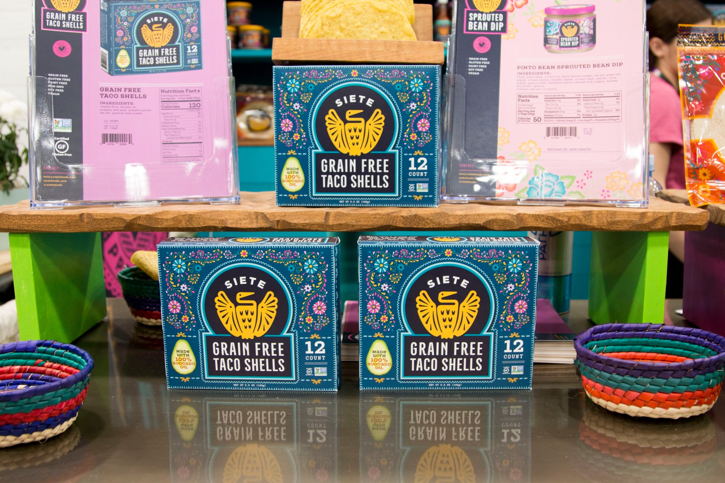 Teal Siete taco shell boxes on a wooden stand with colorful baskets and brochures behind.