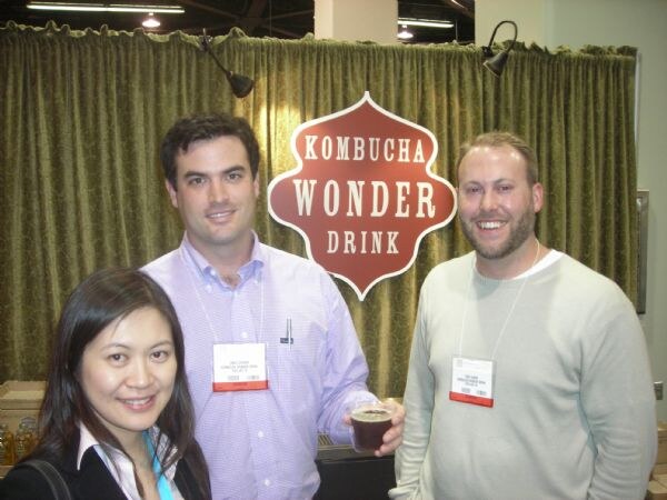 Two men and a woman pose in front of a Kombucha Wonder Drink sign on a green curtain. One man holds a small cup of dark drink, and all three wear badges.