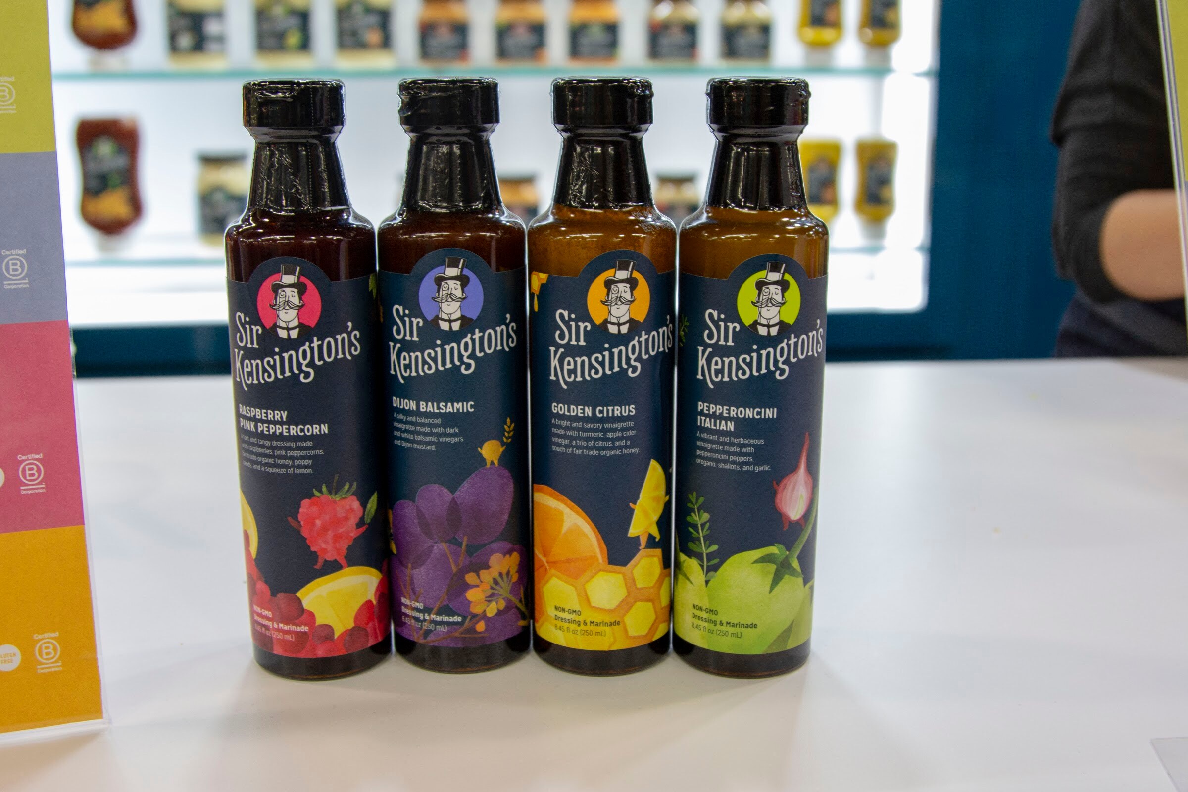 Four dark bottles with colorful Sir Kensington's labels on a white counter with a shelf of bottles behind.