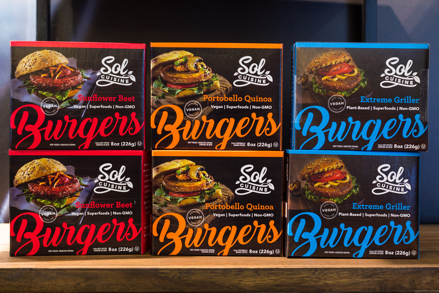 Six black Sol Cuisine burger boxes with red, orange and blue lettering stacked on a wooden shelf.