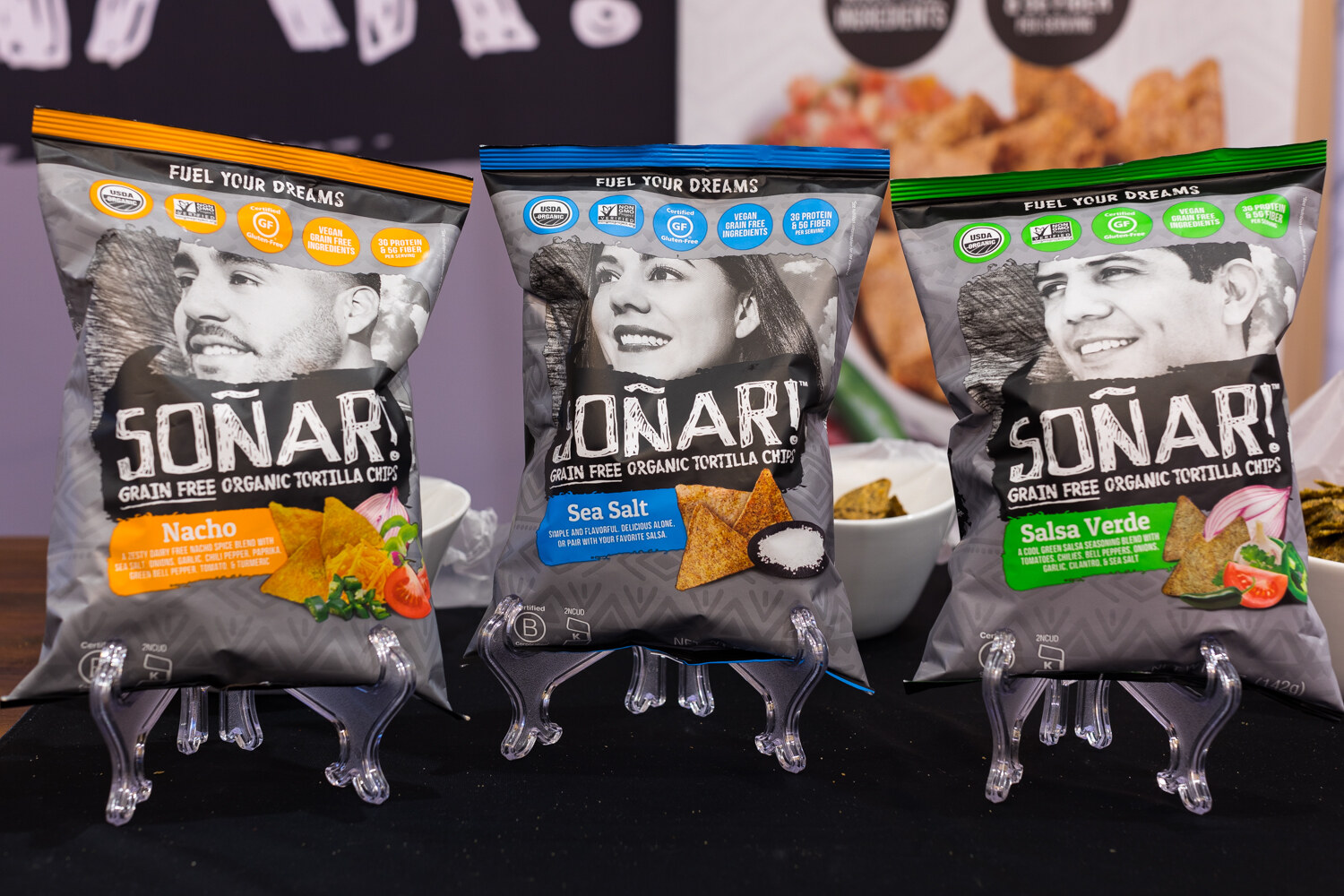 Three gray Sonar chip bags with black and white portraits on clear stands on a black table.